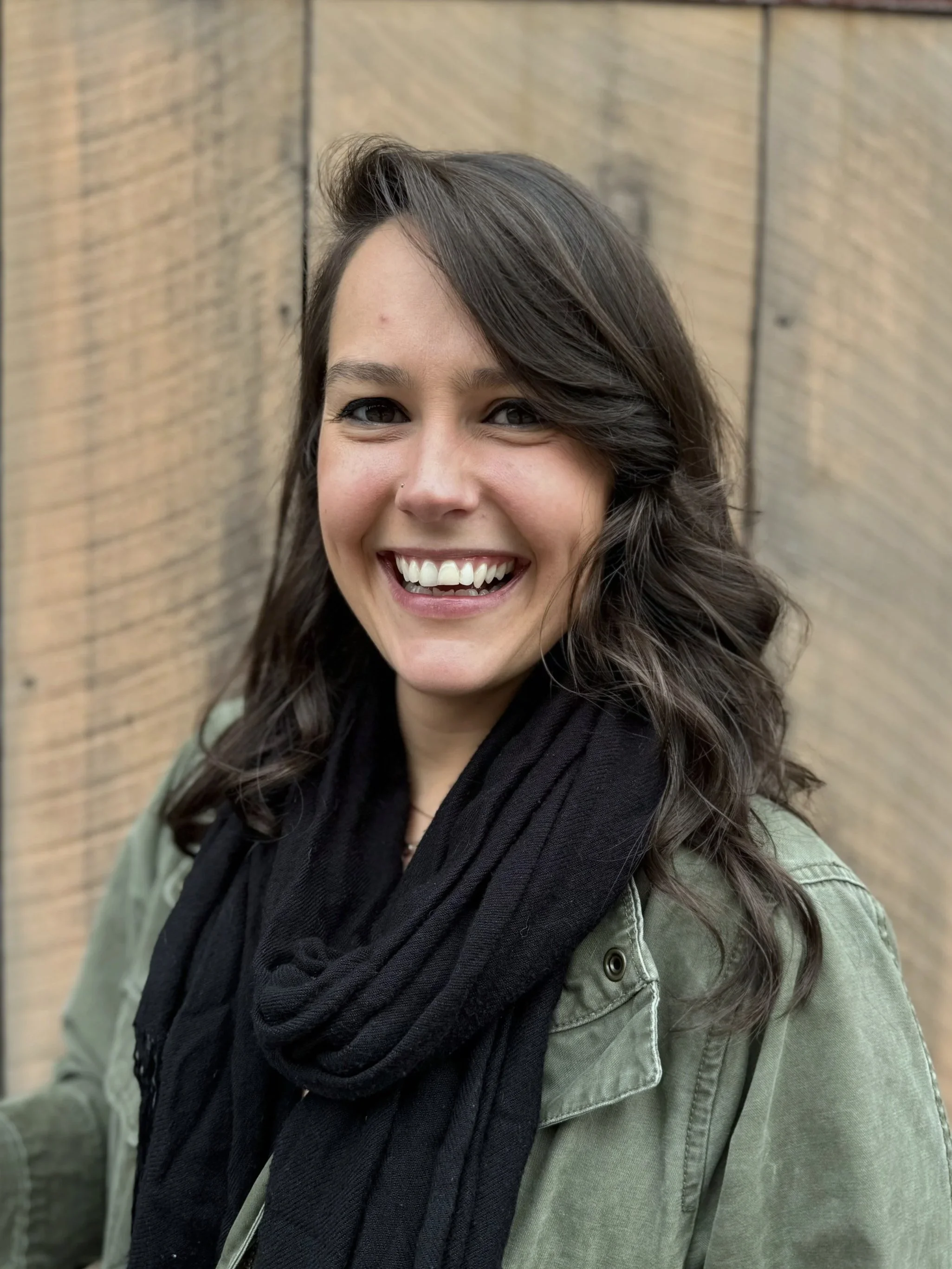 A woman smiling and looking at the camera, with dark brown hair, wearing a black scarf and green jacket, standing in front of a wooden wall.