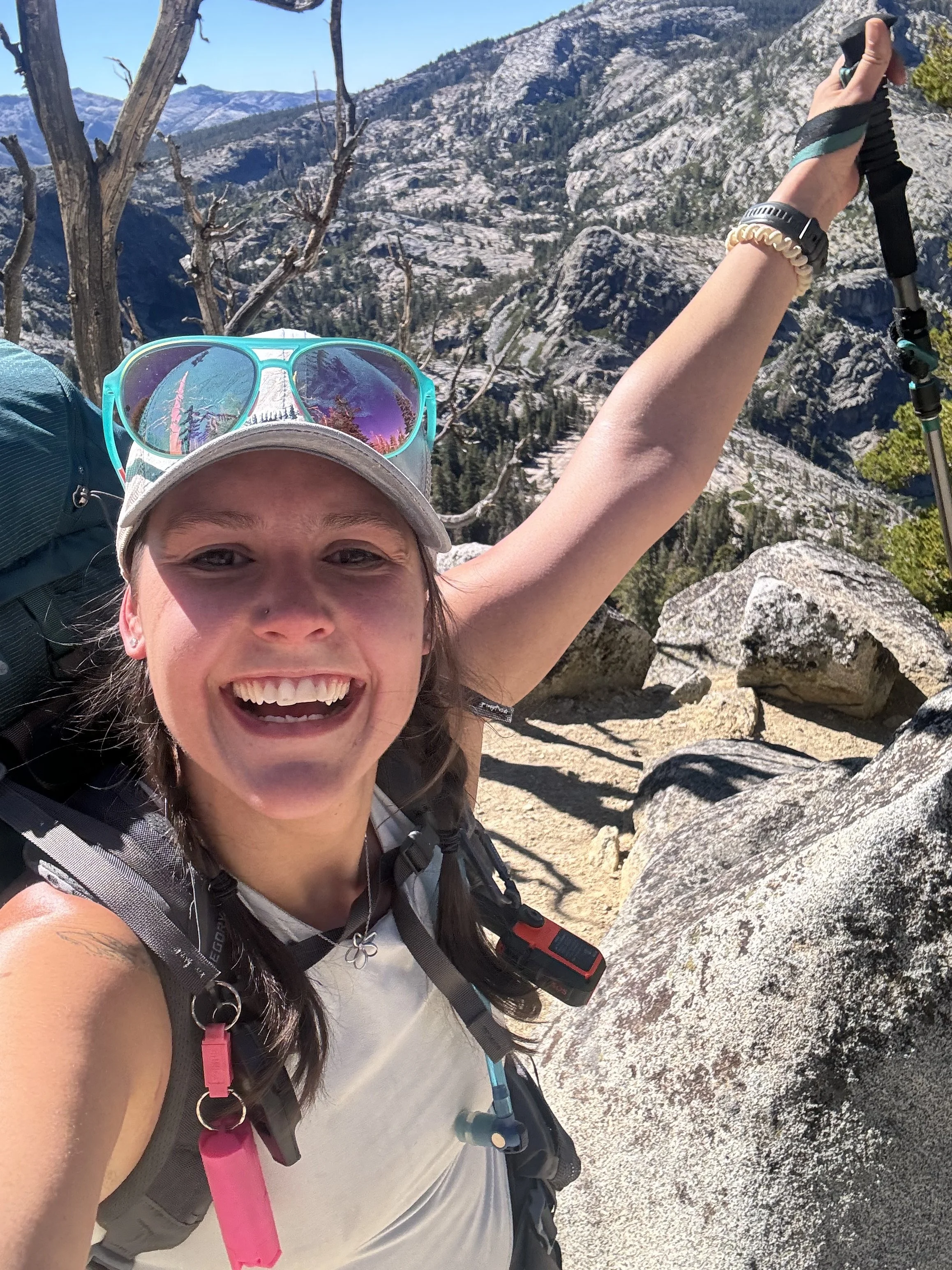 A woman smiling and taking a selfie during a mountain hike, holding hiking pole, wearing sunglasses on her hat, with rocky mountain terrain and dry trees in the background.