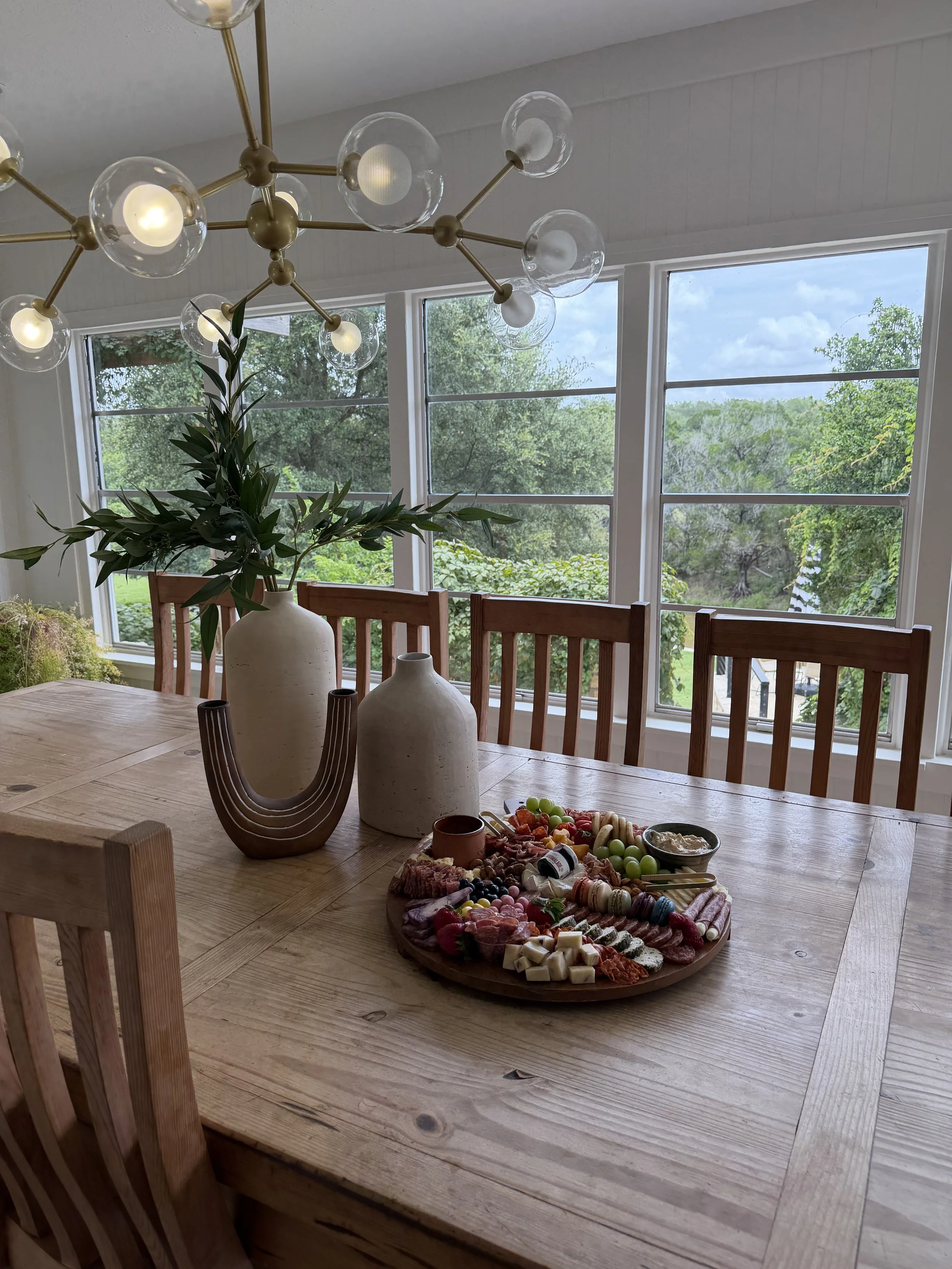 A wooden dining table with a large platter of assorted cheeses, crackers, and fruits, surrounded by wooden chairs. On the table are two large ceramic vases and a decorative wooden sculpture. In the background, large windows reveal a lush green outdoor scene, and a modern chandelier with globe light bulbs hangs from the ceiling.