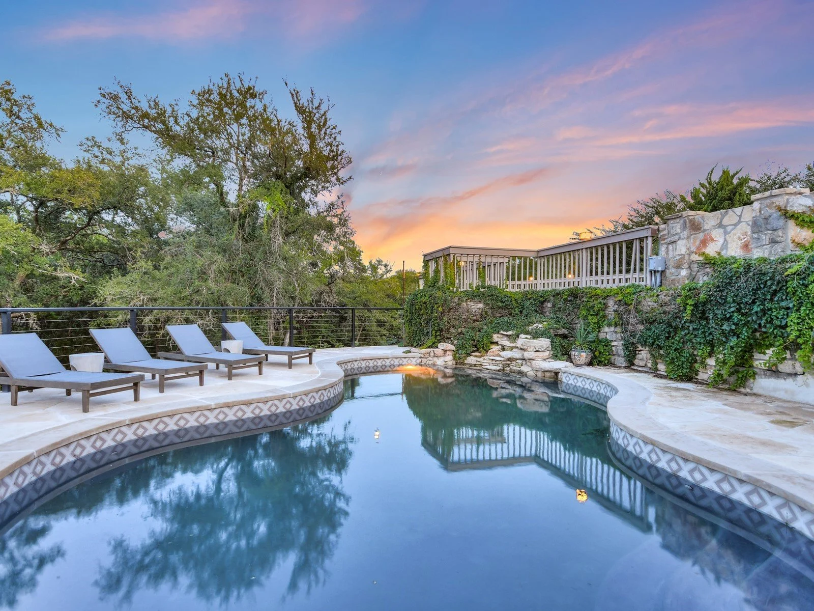 A backyard swimming pool area at sunset with four lounge chairs, lush green trees, and a stone and wood fence, reflecting the colorful sky.