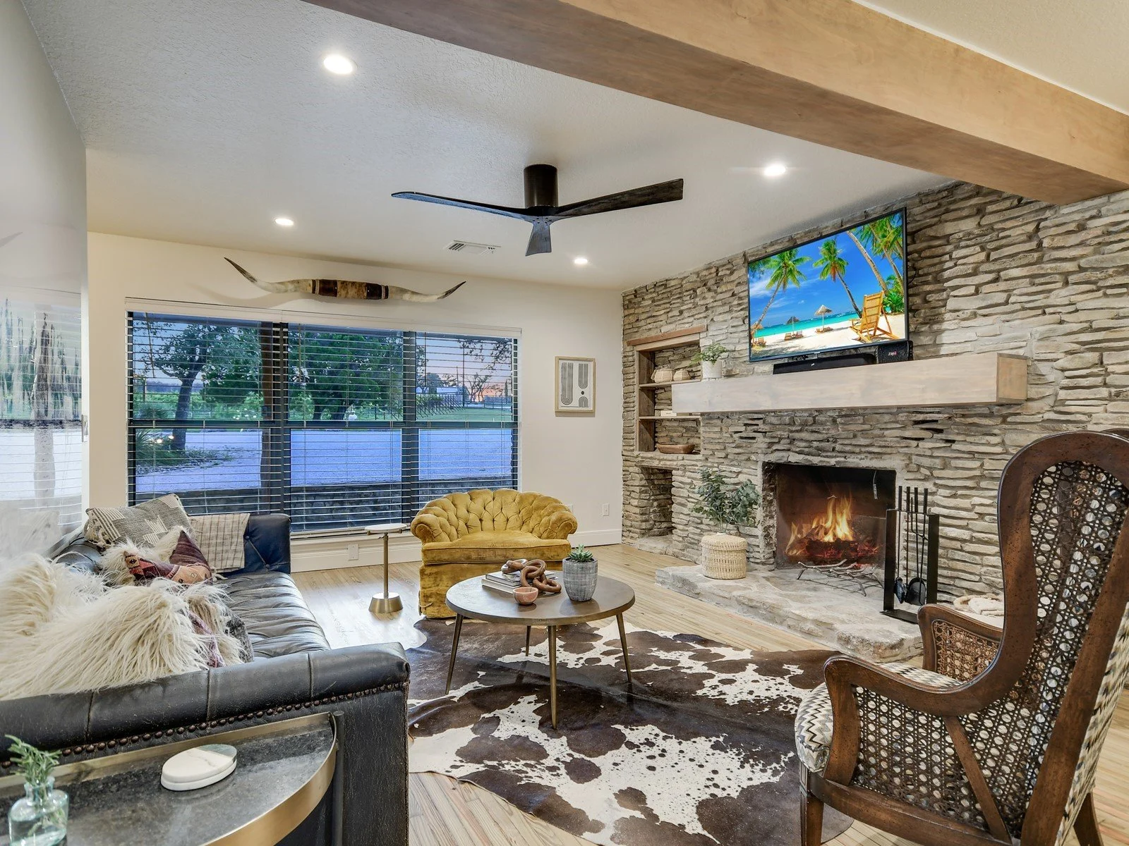 Living room with large window, stone fireplace with fire, mounted TV showing beach scene, yellow velvet armchair, leather sofa with pillows, cowhide rug, wooden side table with decorative items, and a cow skull mounted on the wall.