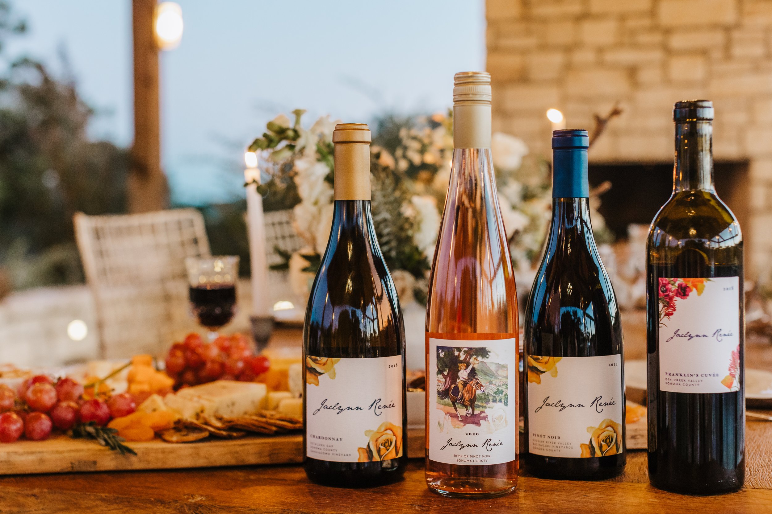 A close-up of four wine bottles with labels, set on a wooden table at a gathering. The background includes blurred flowers, a brick wall, and food items such as grapes and cheese.