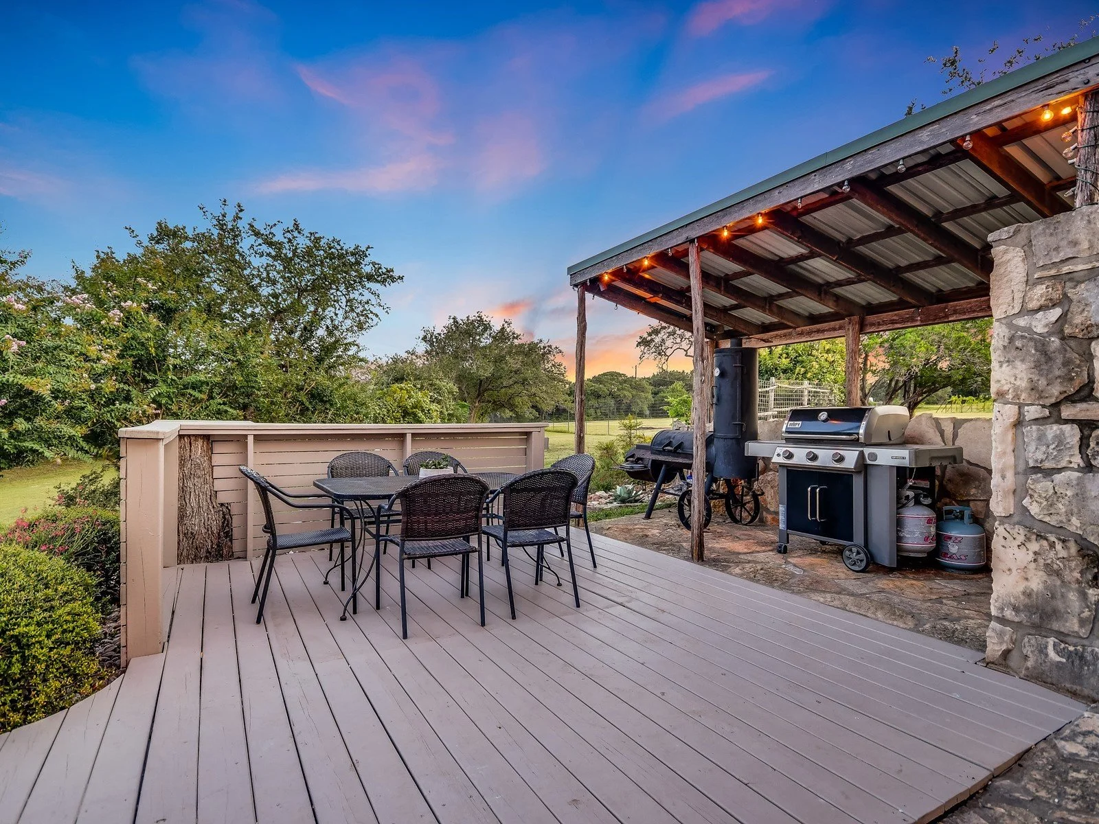 Outdoor patio with a dining table and six chairs, a covered area with a grill, a stone wall, and a view of trees and a field at sunset.