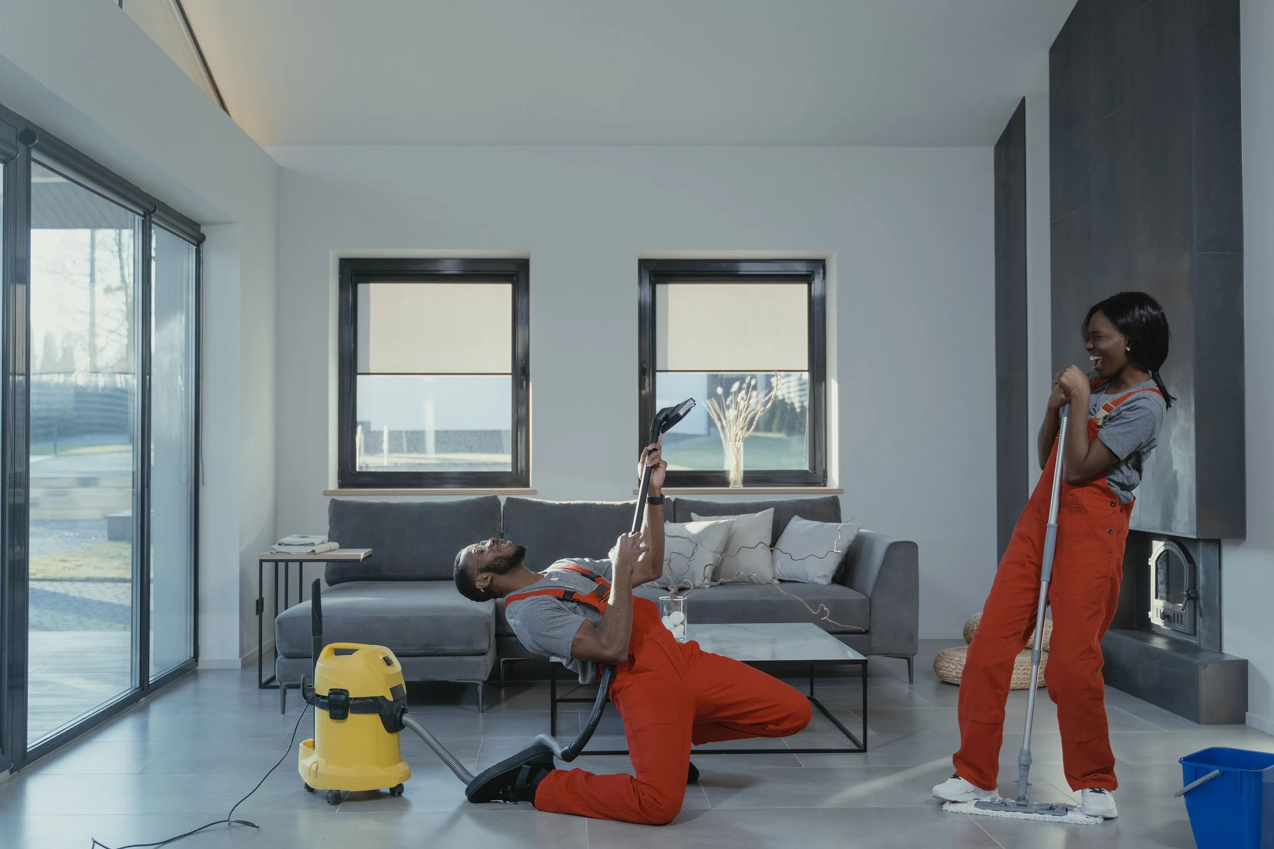 Two cleaning workers in orange overalls are enjoying a moment of laughter in a modern living room. One is lying on the floor using a vacuum cleaner, and the other is standing with a mop. The room has large windows, a gray sofa, and a colorful decorative item in the background.