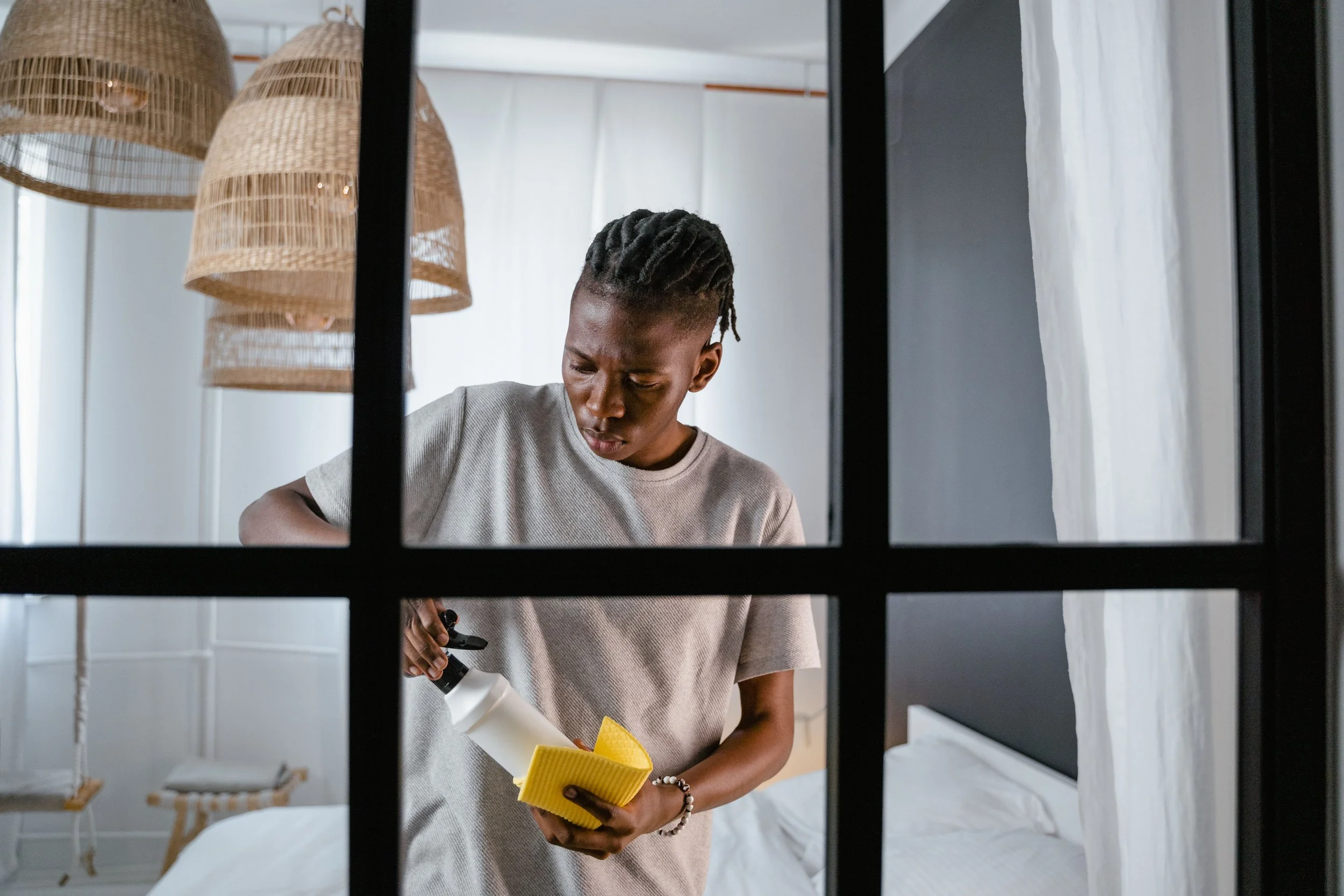 A person with short dreadlocks and a light-colored t-shirt cleaning a window with a yellow sponge and a spray bottle in a modern, minimalistic room with hanging woven lampshades and white curtains.