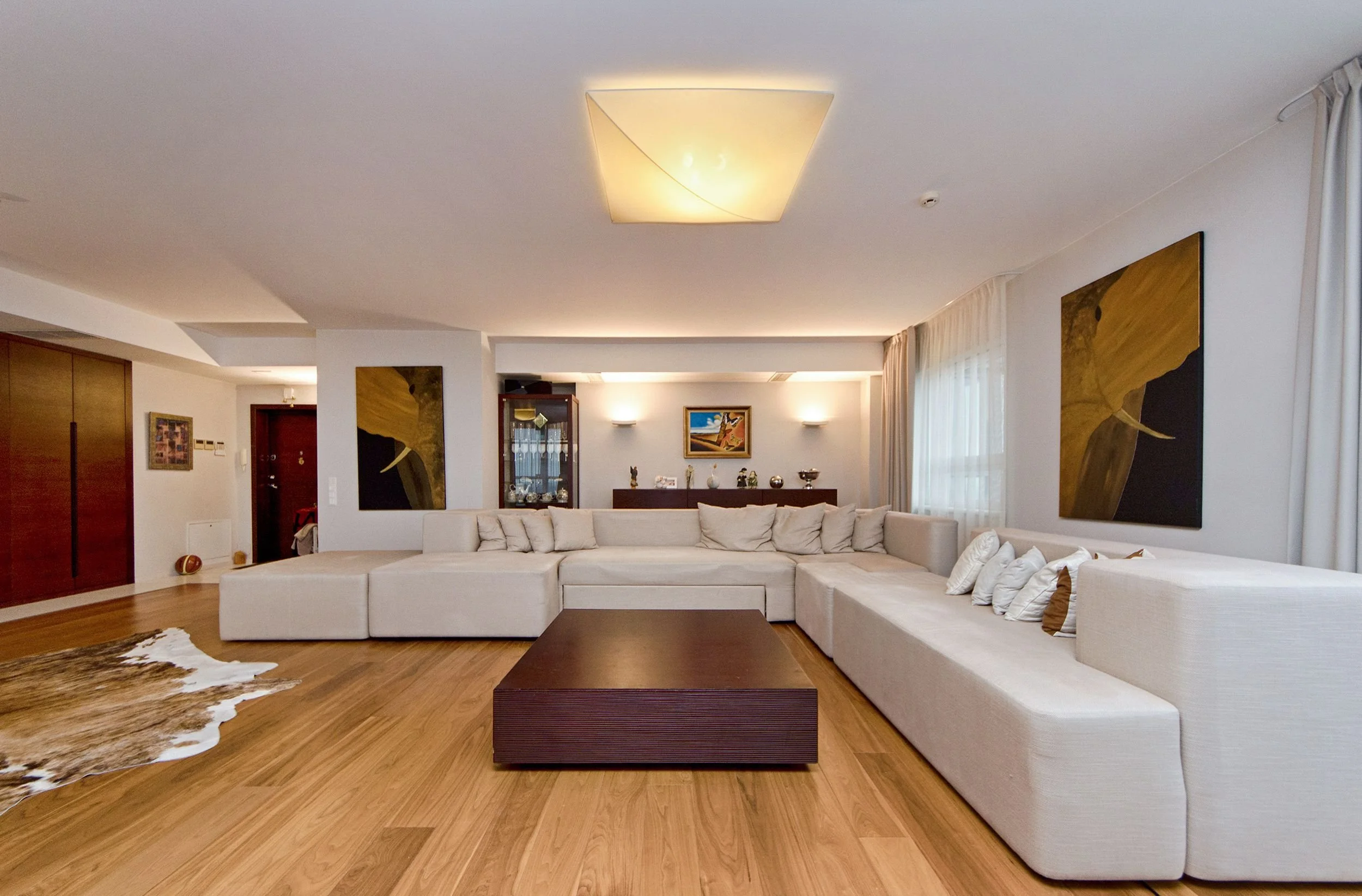 A spacious living room with white sectional sofa, wooden coffee table, abstract artwork, and a cowhide rug on hardwood floor.