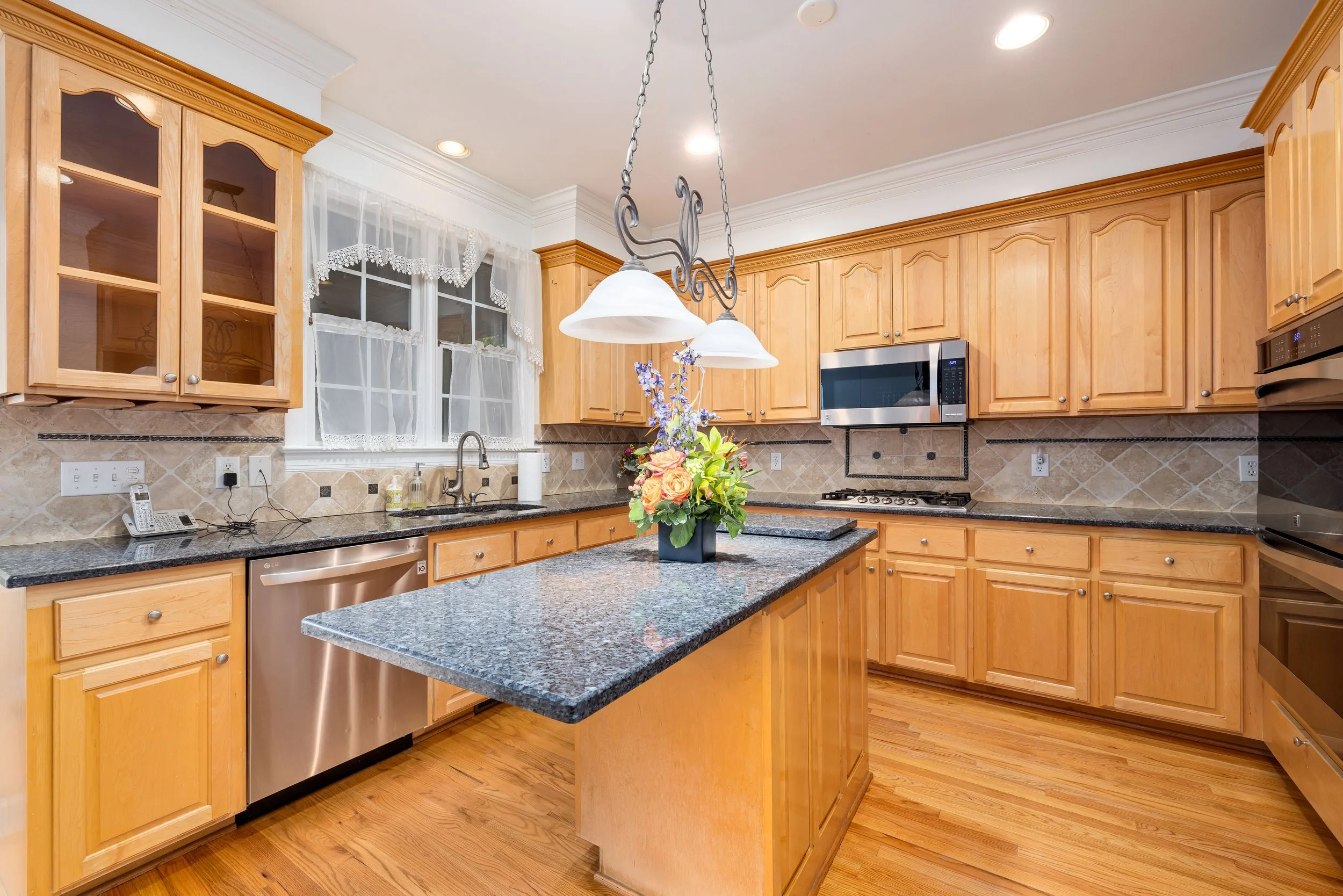 A spacious kitchen with wooden cabinets, granite countertops, a central island with a flower arrangement, and a window with sheer lace curtains.