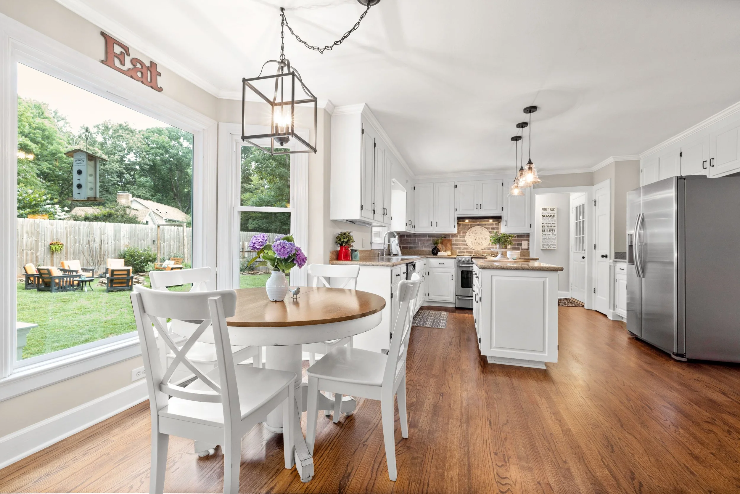 A bright kitchen with white cabinets, a round wooden dining table with white chairs, and large windows overlooking a backyard with outdoor seating. The kitchen has stainless steel appliances, pendant lights, and decorative plants and flowers.