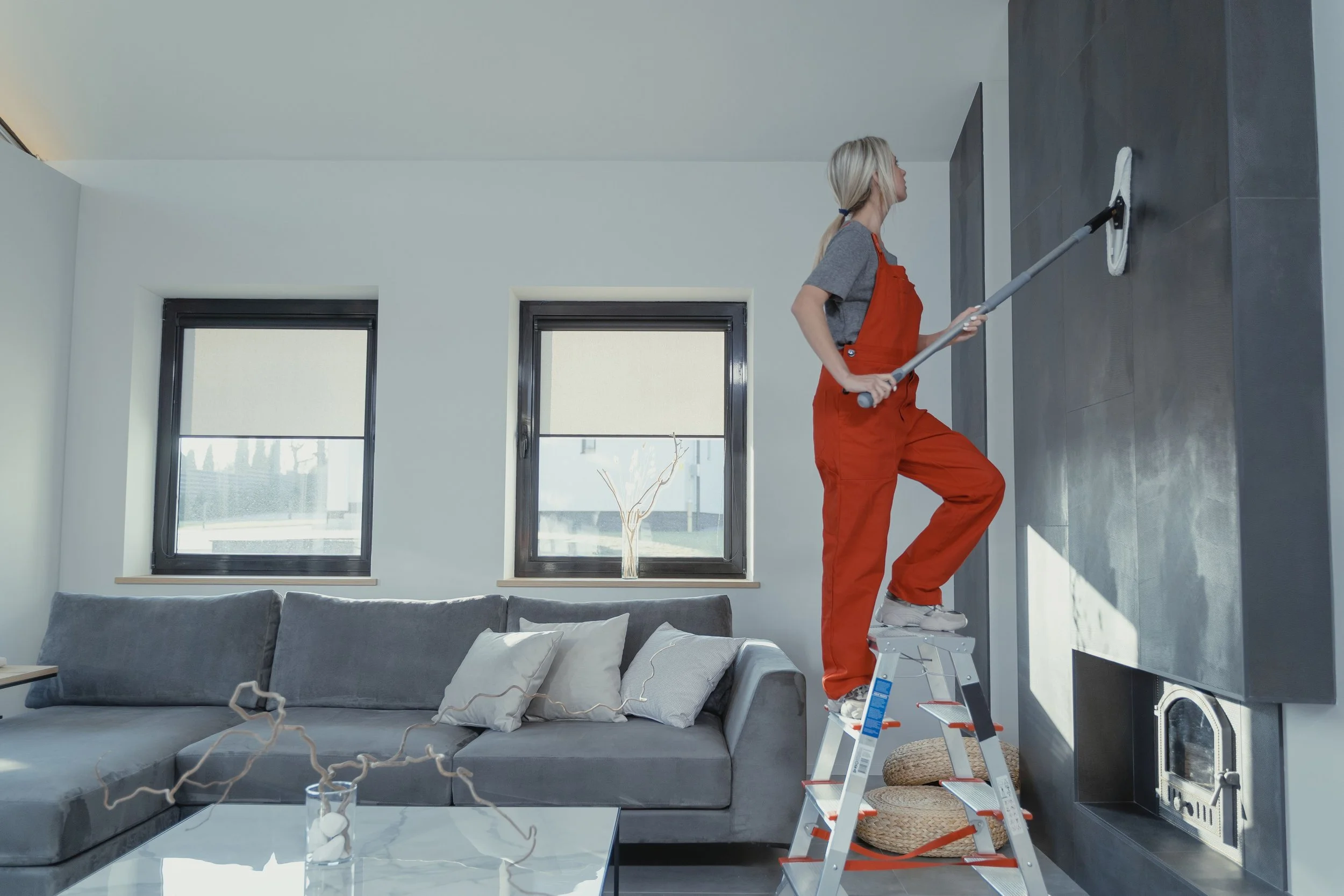 A woman cleaning a dark gray wall in a modern living room with a step ladder, a gray sectional sofa, and large windows.