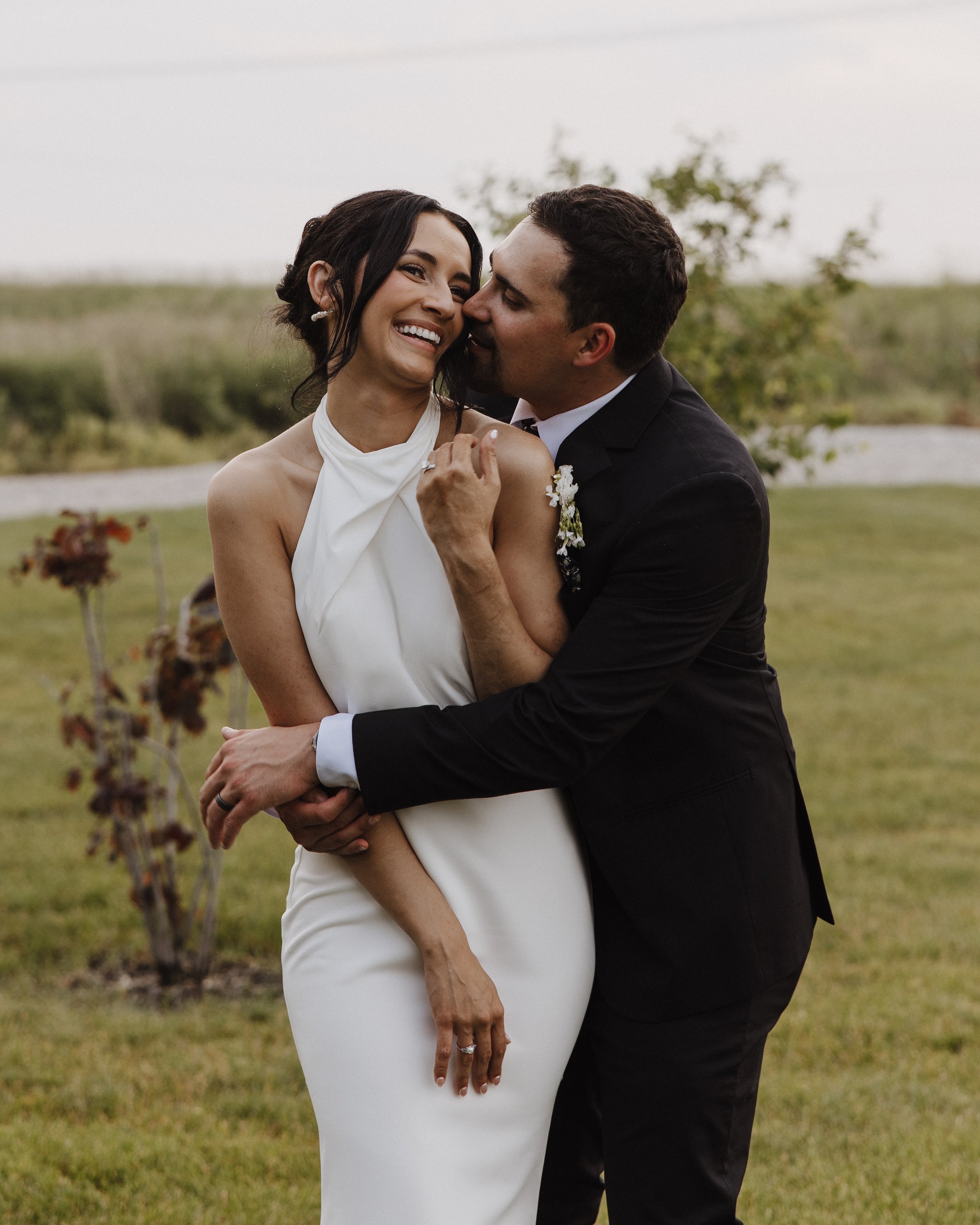 A happy couple embraces outdoors on a grassy field with trees and a pond in the background. The woman wears a white dress and the man wears a black suit. They are smiling and close to each other, sharing a tender moment.
