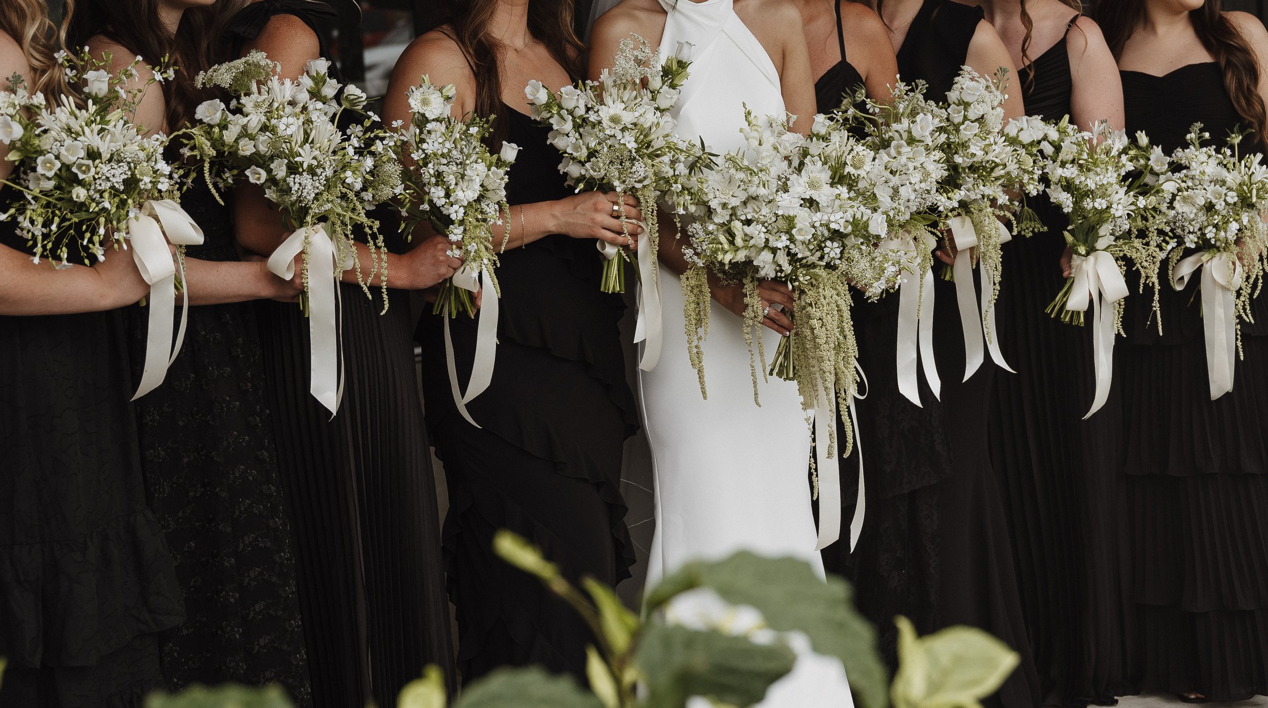 Bridal party holding white floral bouquets at a wedding, with the bride in a white dress and bridesmaids in black dresses.