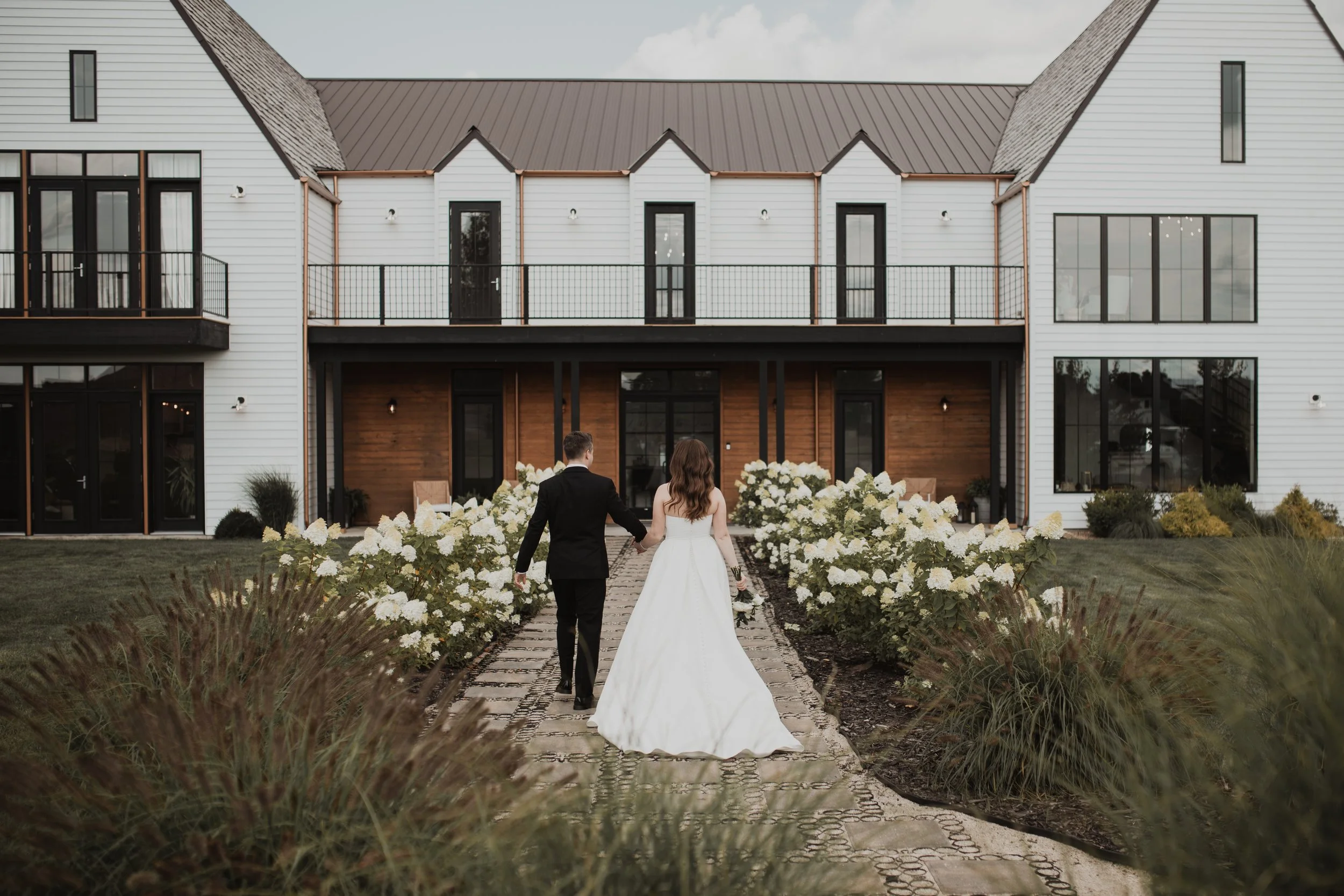 A bride and groom walking hand in hand on a stone path towards a modern white house with a balcony, surrounded by white flowers and greenery.