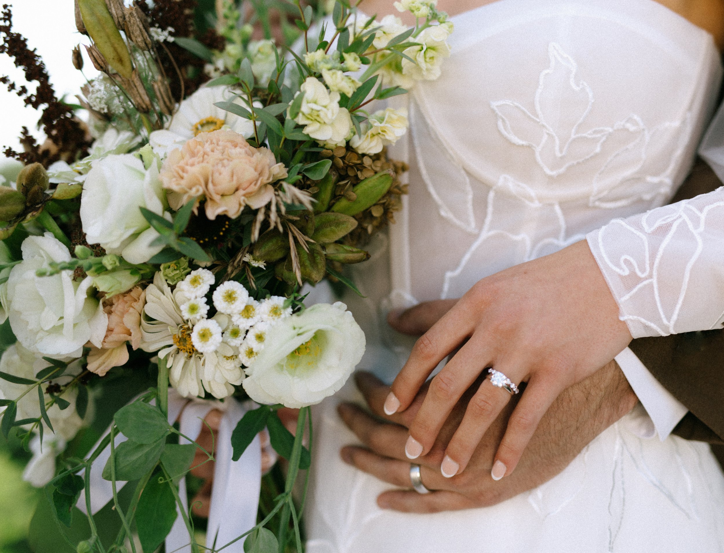 Close-up of a bride and groom's hands joined, showing wedding rings, against a bouquet of white and green flowers and white fabric with embroidery.