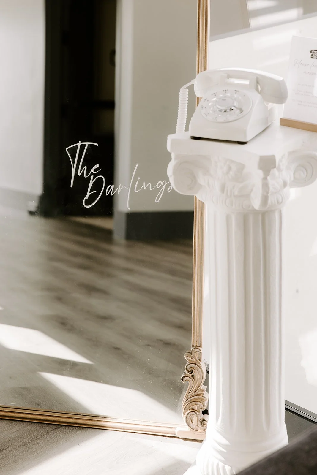 A white vintage rotary phone on a decorative white pedestal next to a mirror with the words "The Darling" written on it, and a framed paper in the background.