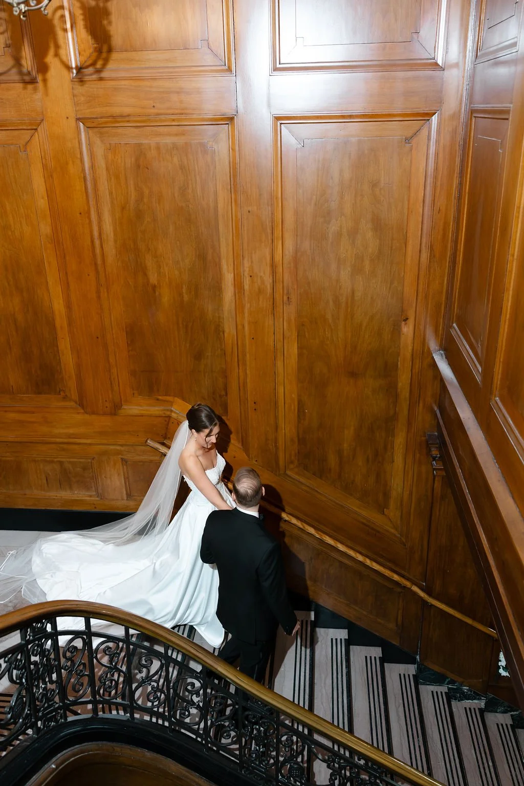 Bride in a white wedding dress and veil walking down a staircase with a man in a black suit, inside a building with wooden paneling and black and white striped flooring.