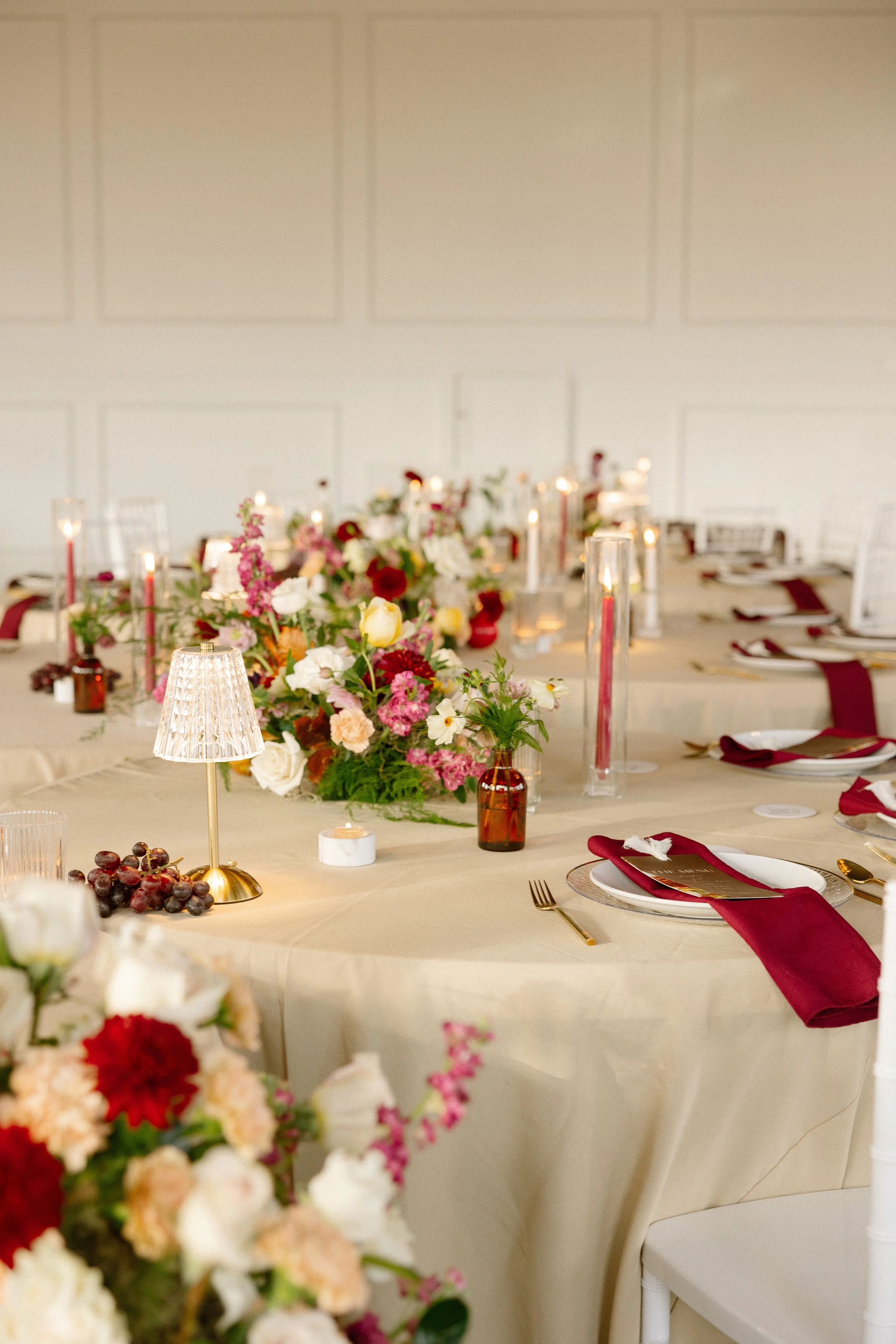 A beautifully decorated dining table with a floral centerpiece, candles, plates, and burgundy napkins, set for a formal event in a room with white walls.