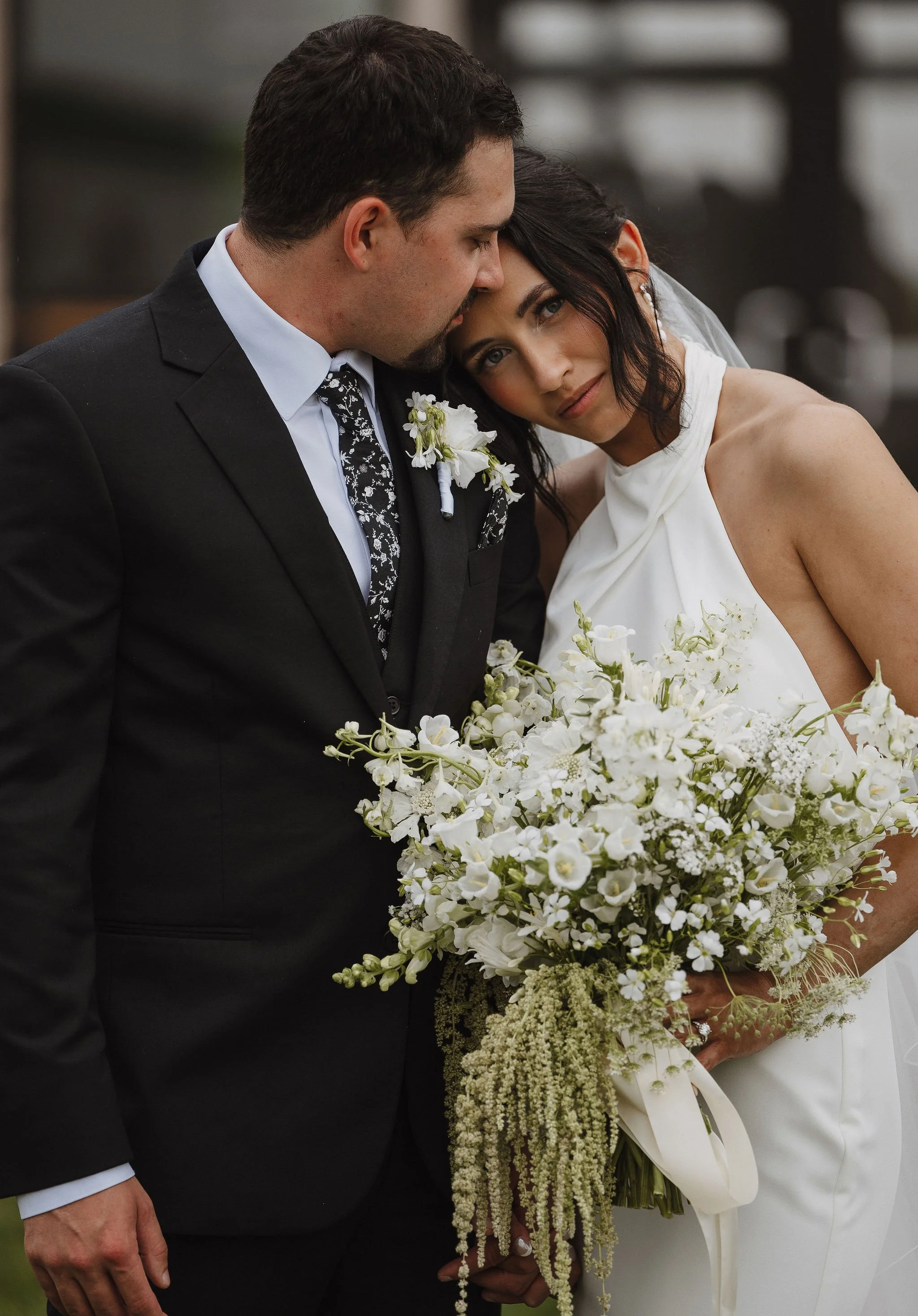 A bride and groom at their wedding, the groom is wearing a black suit and tie with a white shirt and boutonnière, the bride is wearing a white wedding dress holding a large bouquet of white flowers, and she has floral earrings with a veil.