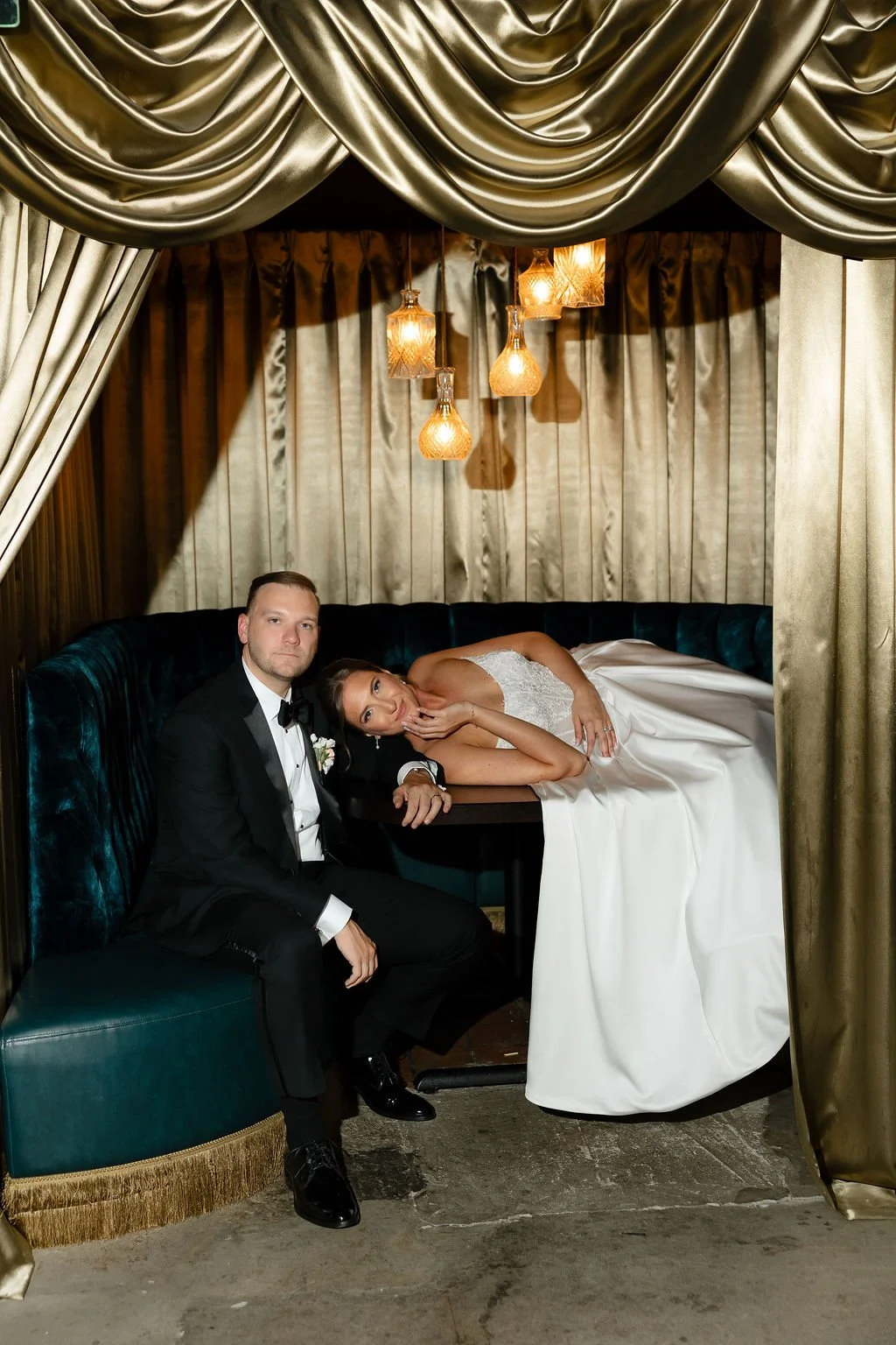 A bride and groom in wedding attire sitting inside a luxurious, gold and velvet draped booth. The bride is lying on the table with her head resting on the groom's shoulder, while he sits next to her, looking at the camera. The booth features elegant 
