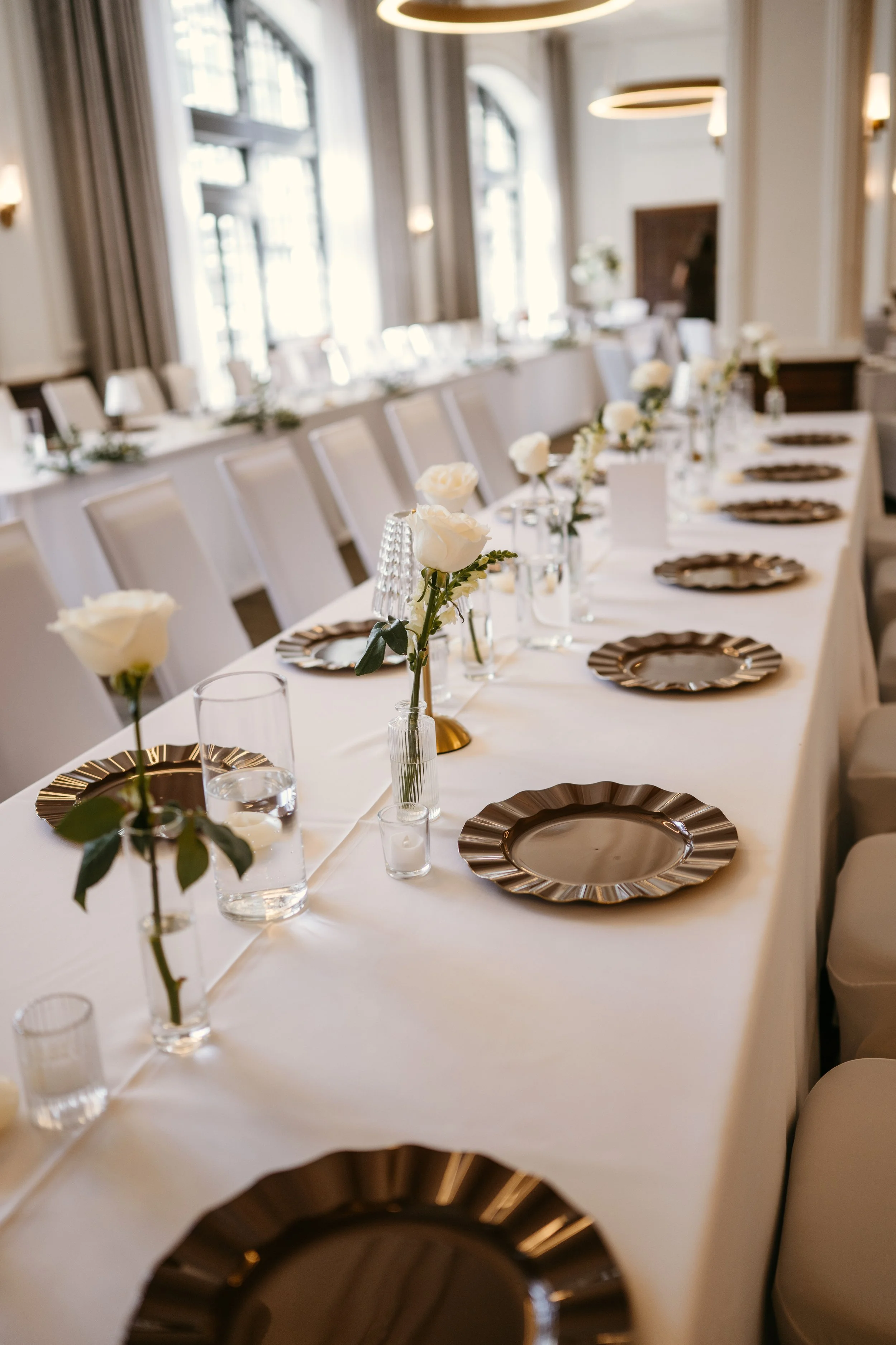 Elegant dining room with long table covered in white tablecloth, set with gold-rimmed plates, clear glasses, and small floral centerpieces with white roses in glass vases. Soft lighting and large windows with drapes create a bright atmosphere.
