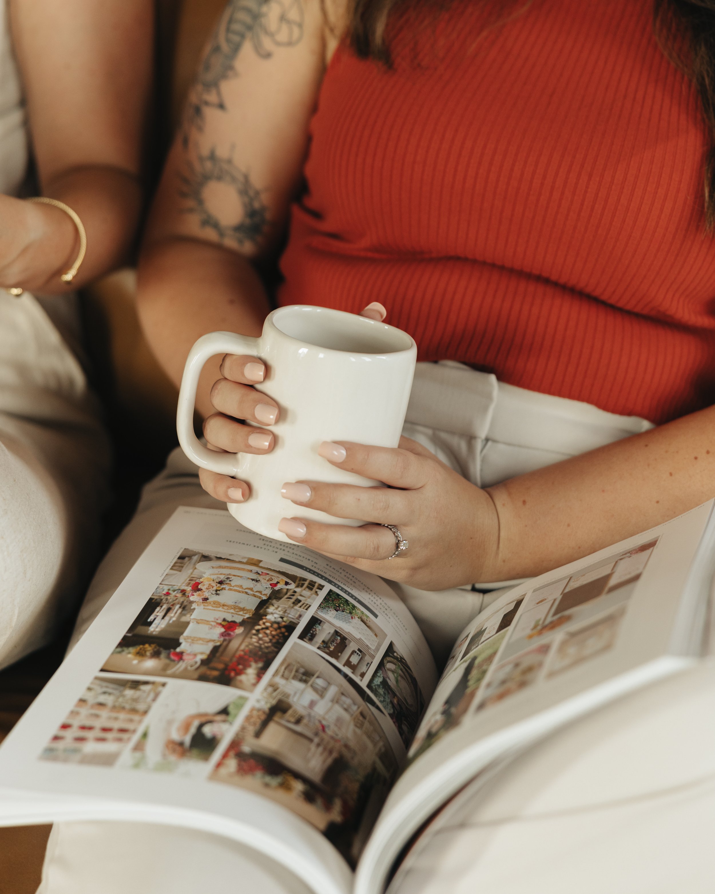 A person holding a white ceramic mug while looking at a large catalog or magazine with interior design images.