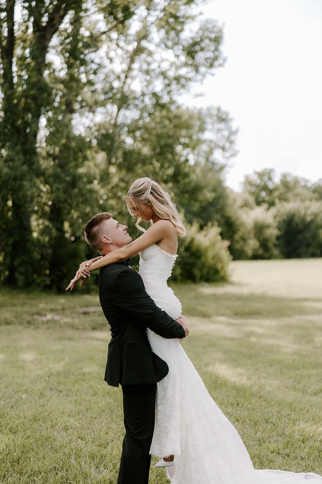 A bride in a white wedding dress being lifted by a groom in a black tuxedo outdoors on a grassy field with trees in the background.
