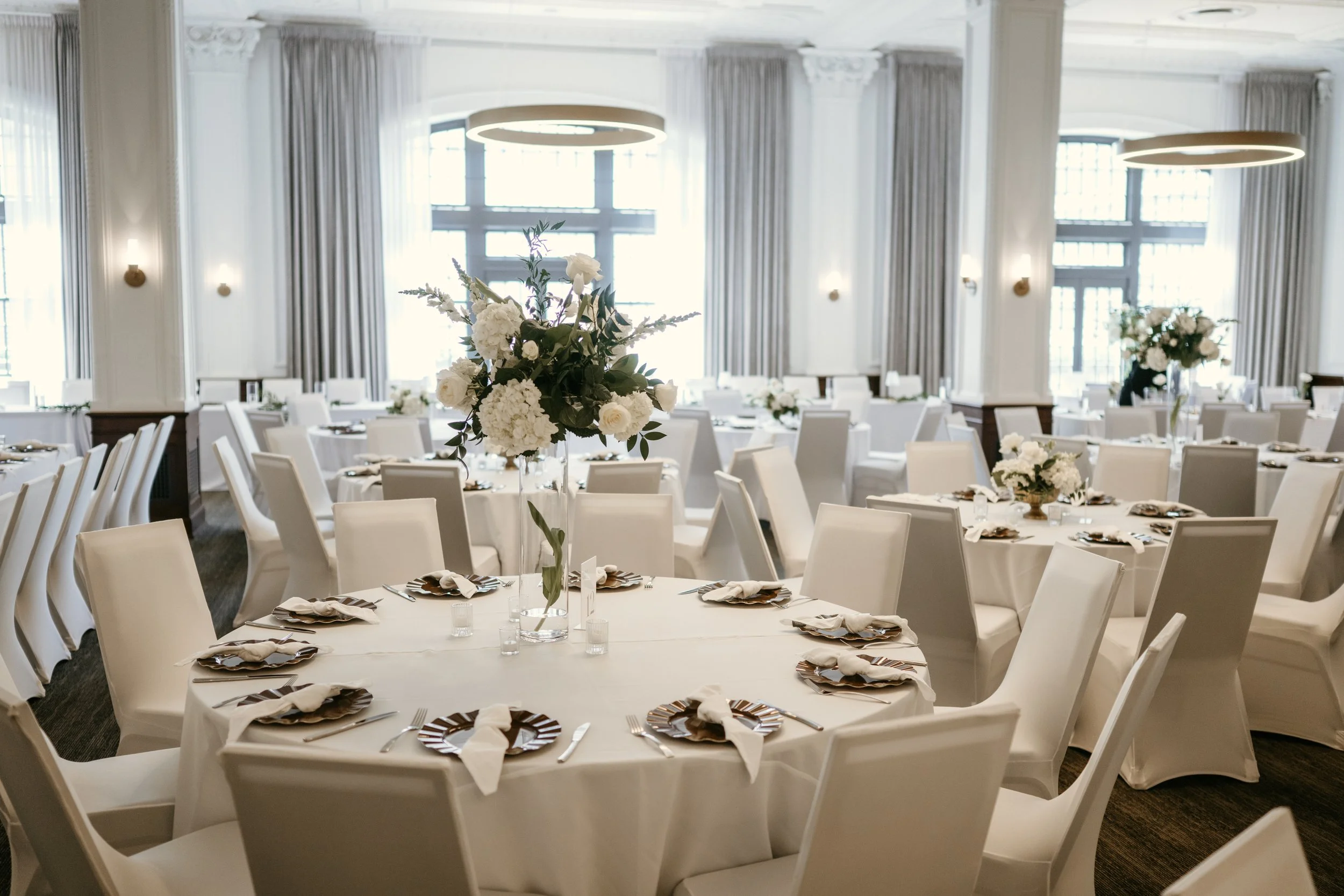 A banquet hall set up for a formal event with round tables, white chairs, and floral centerpieces, featuring high ceilings and large windows with curtains.