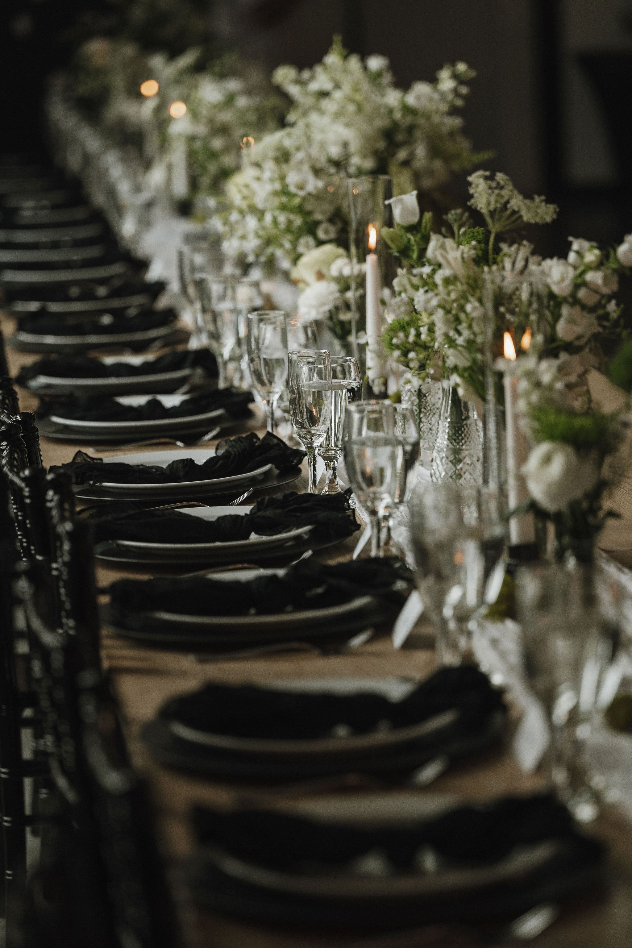 A long dining table set with black napkins, glassware, and floral centerpieces, with candles, in a dimly lit room for a formal event or celebration.