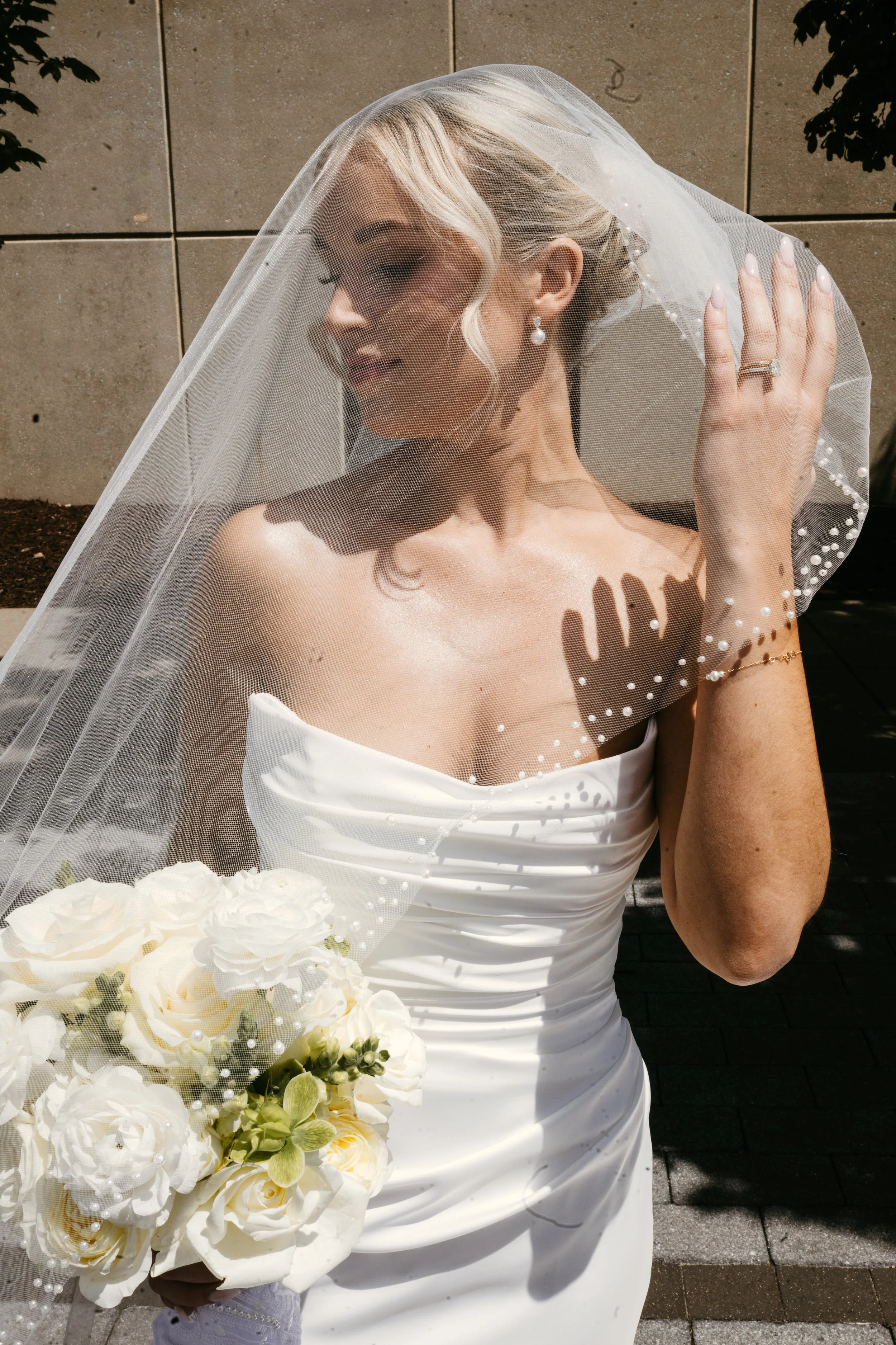 A woman in a white wedding dress with a veil holding a bouquet of white flowers, standing outdoors against a concrete wall.