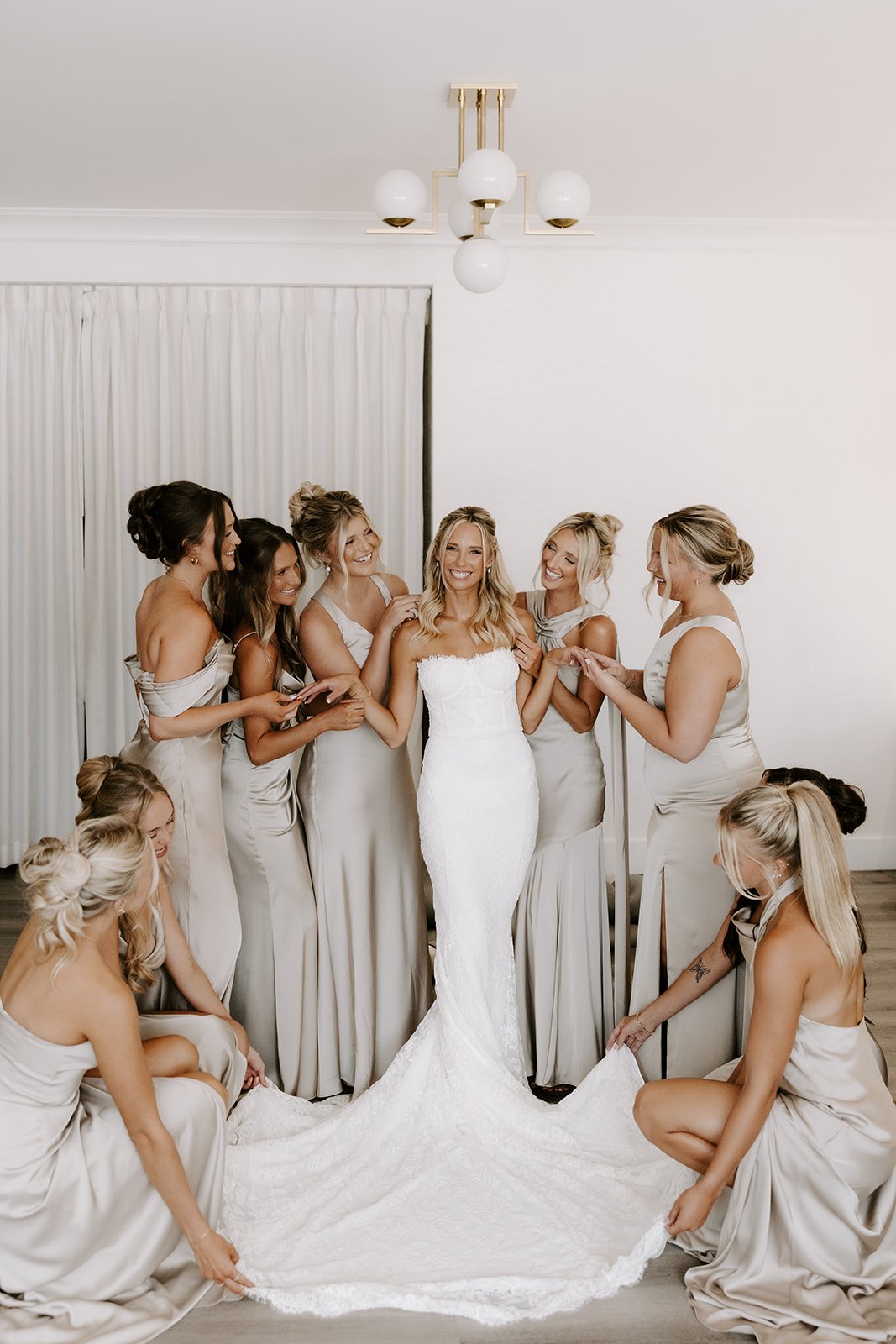 Bride in white wedding dress surrounded by bridesmaids in beige dresses, in a room with white walls and ceiling light fixture.