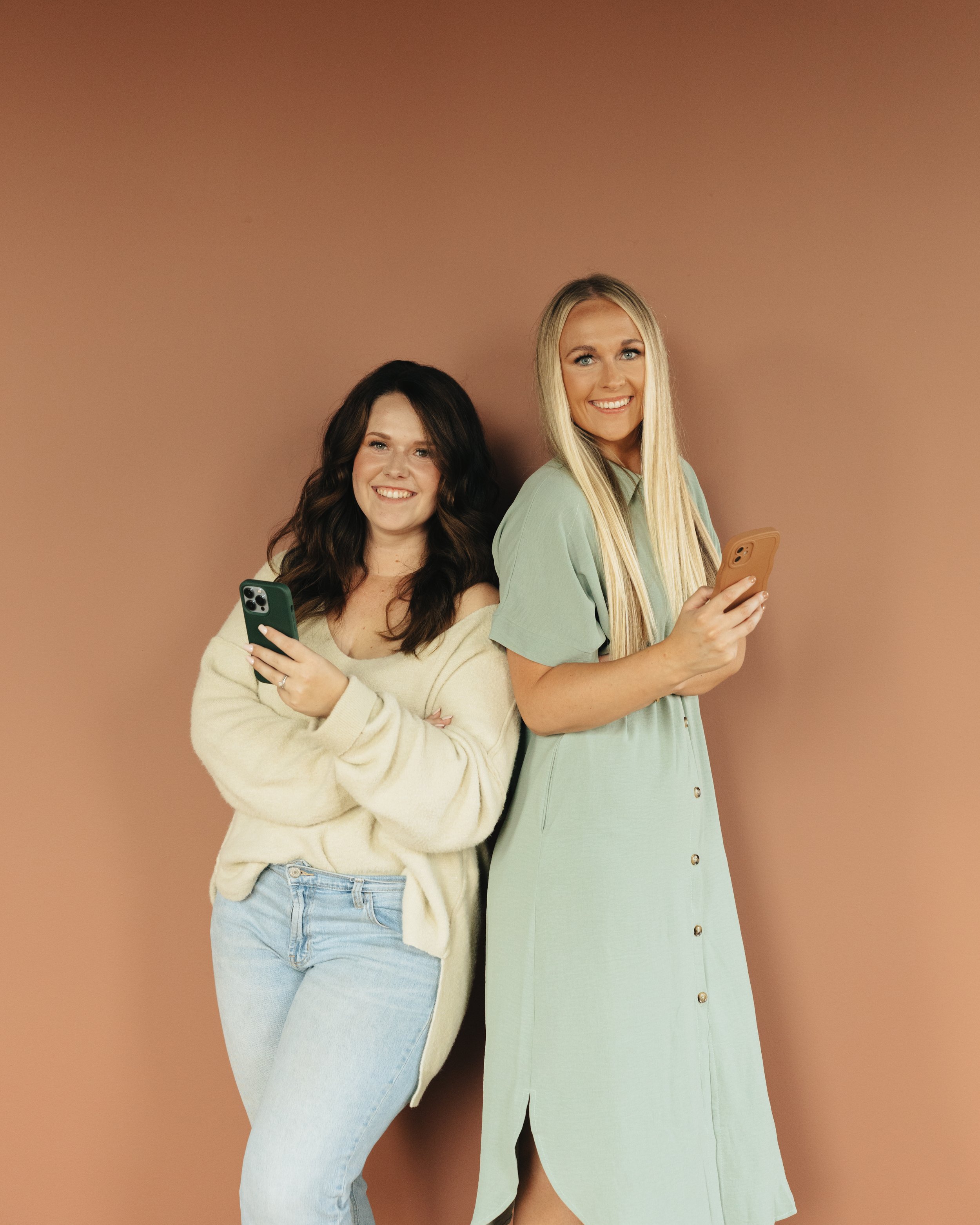 Two women standing against a plain brown wall, holding phones and smiling, with one woman in a beige sweater and jeans, and the other in a light green dress.