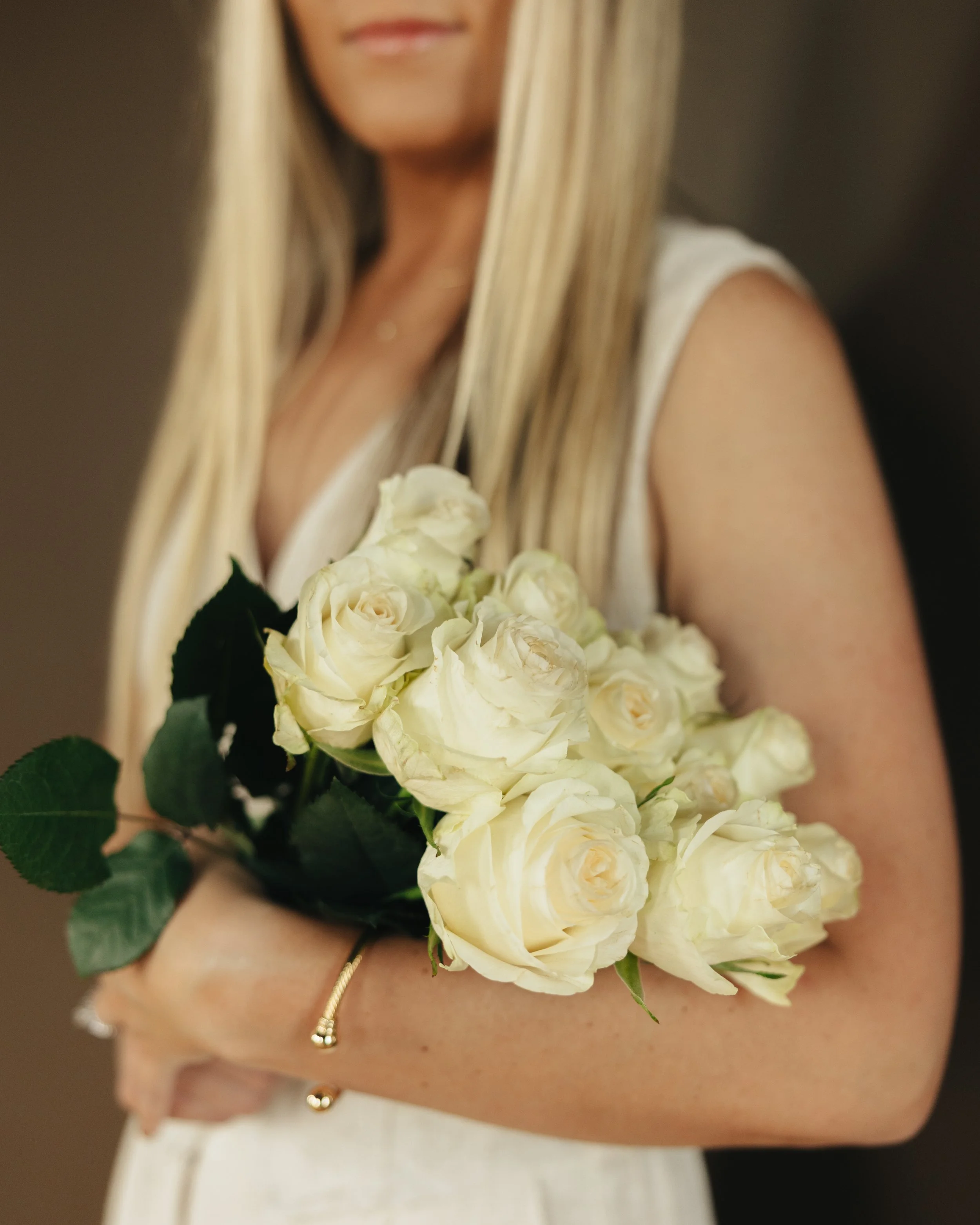 A woman in a white dress holding a bouquet of white roses with green leaves, with only part of her face and upper body visible.