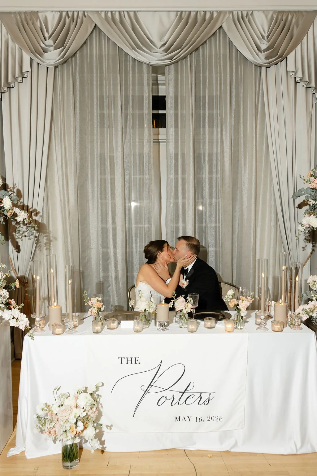 A bride and groom sharing a kiss at their wedding reception table, decorated with flowers and candles, with a sheer curtain and draped valance behind them.