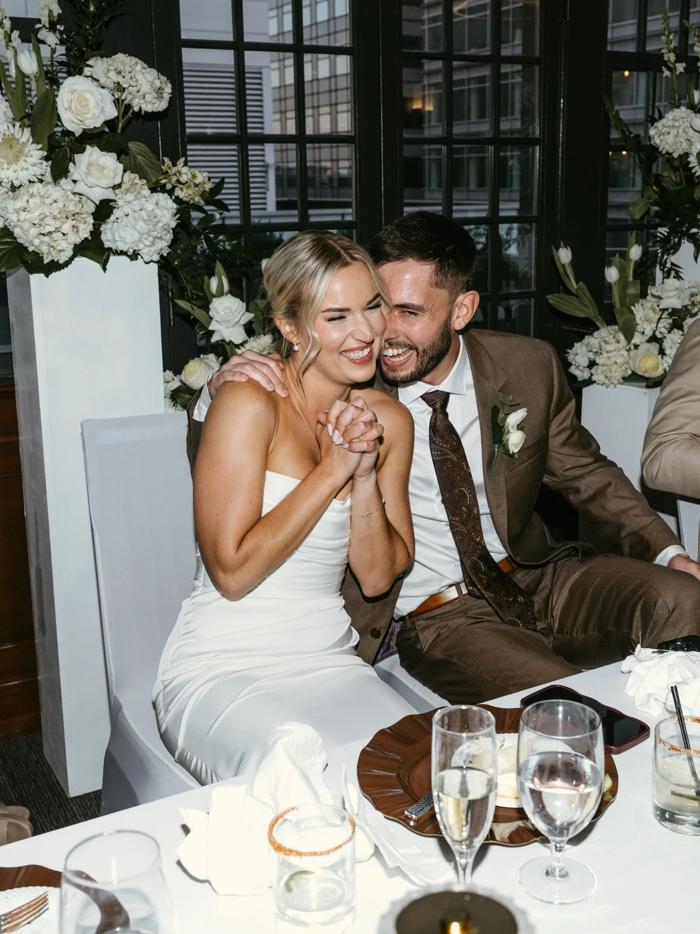 A happy bride and groom sitting at a wedding reception table, laughing and sharing a joyful moment together. The bride is in a white wedding dress, and the groom is dressed in a suit with a tie. The table is decorated with glasses, plates, and floral arrangements, and there are large windows and floral decorations in the background.