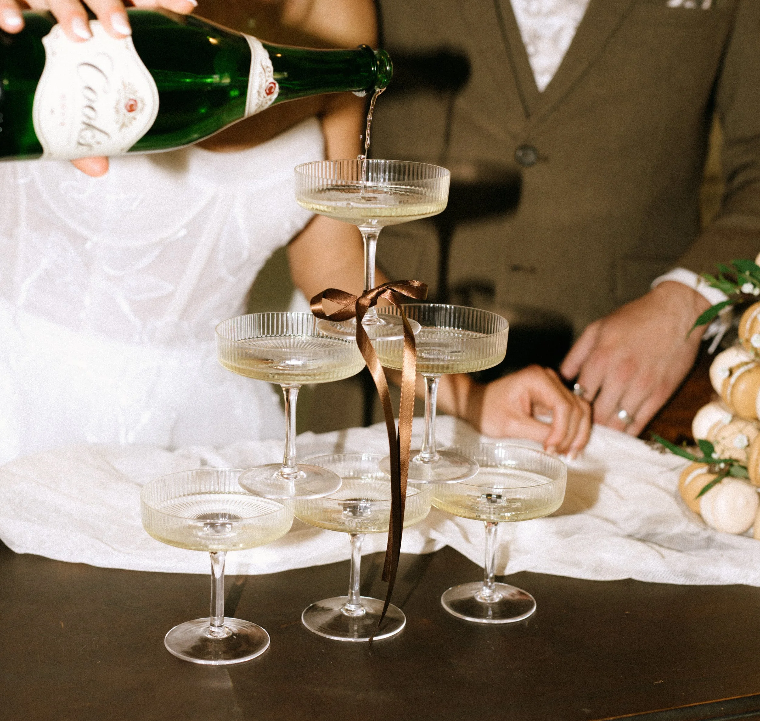 A hand pouring champagne into a stack of crystal champagne glasses tied with a brown ribbon, with a person in a brown suit in the background.