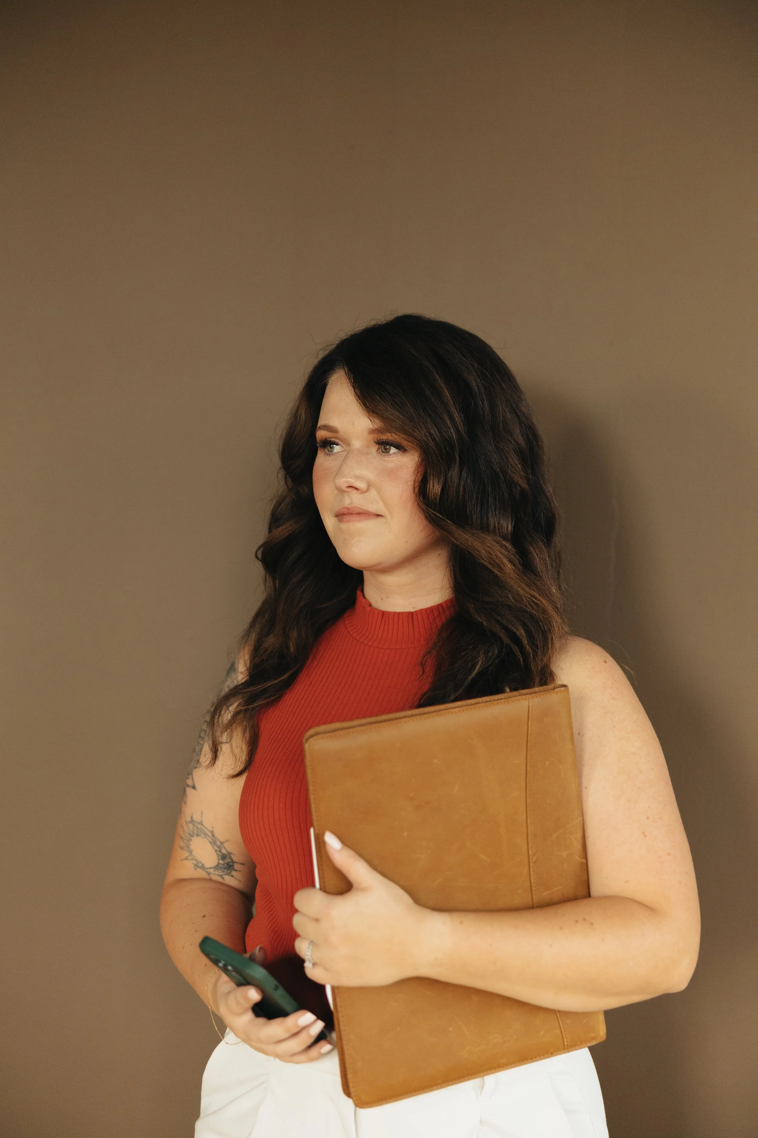 A woman with wavy dark brown hair, wearing a red sleeveless top, holding a brown notebook and a smartphone, standing against a plain beige background.