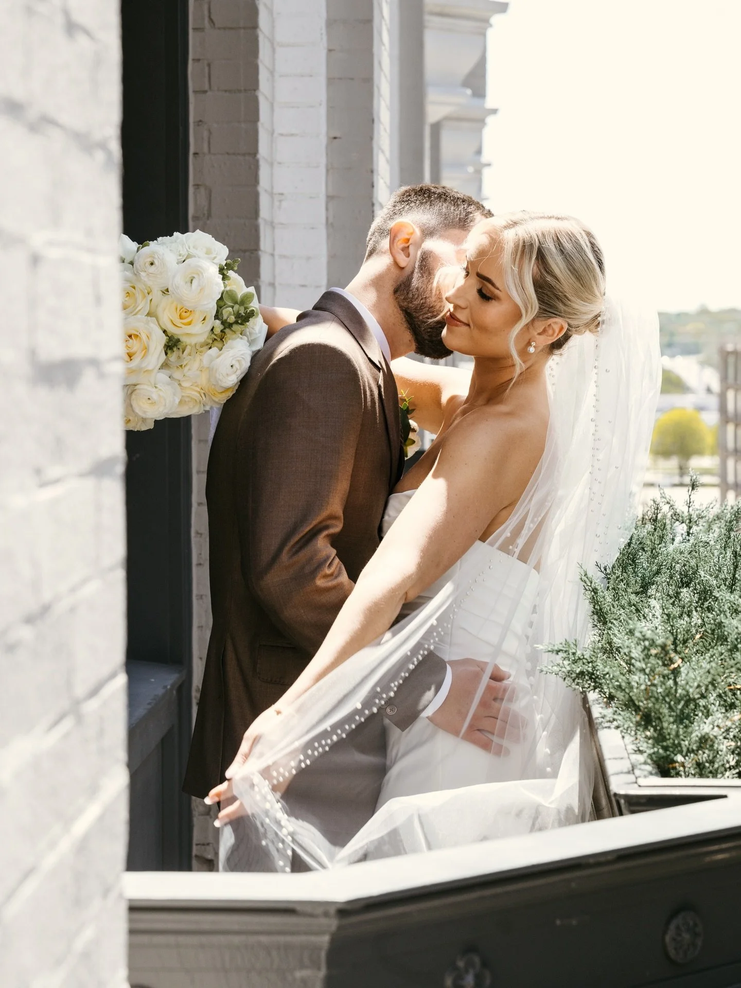 A bride and groom sharing a kiss on a balcony, with the bride wearing a veil and pearl earrings, and the groom in a dark suit. They are standing close to a window with a bouquet of white roses nearby, and outdoor scenery is visible in the background.
