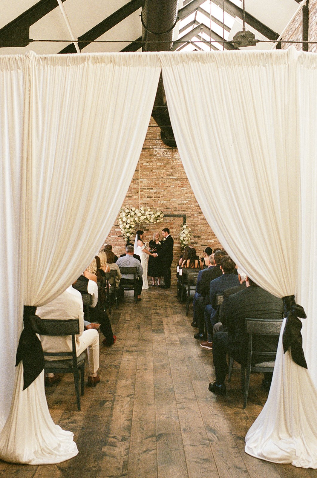 A wedding ceremony taking place inside a venue with exposed brick walls and high ceilings. The bride and groom stand facing each other under a floral arch with white flowers, with an officiant between them. Guests are seated on either side of the aisle, watching the ceremony.