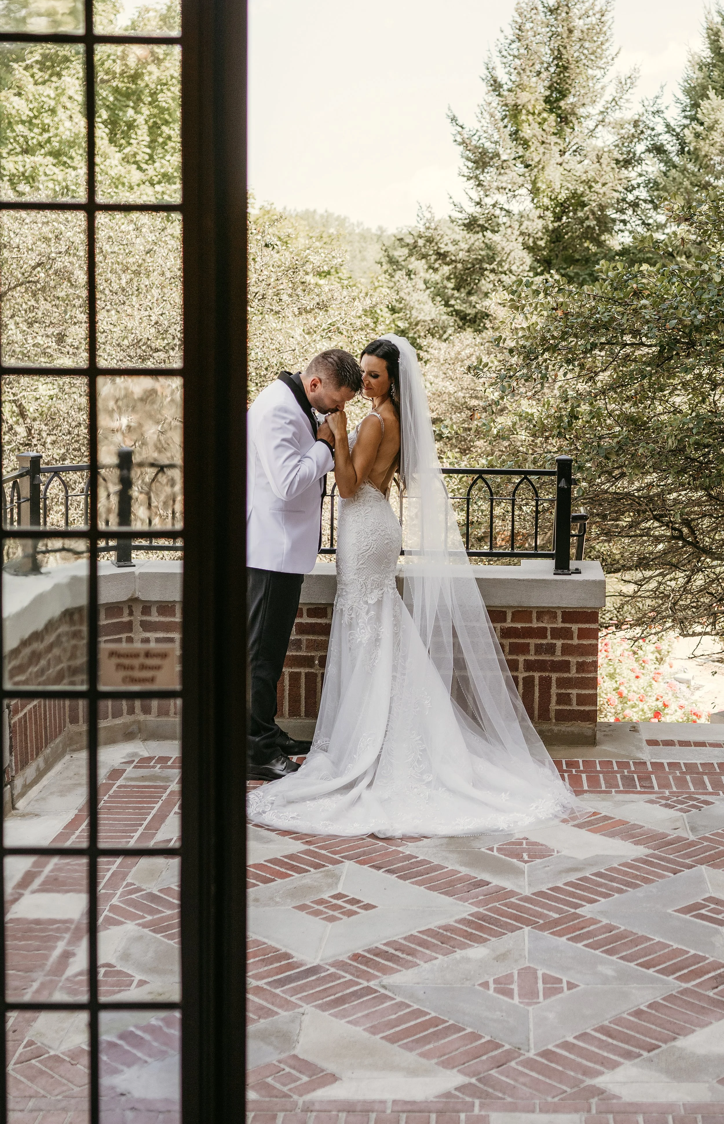 A bride and groom stand close together on a brick and stone patio, framed by a doorway, with green trees in the background. The groom, wearing a white tuxedo jacket, is holding the bride's hands and kissing them. The bride is in a fitted lace wedding gown with a long veil, looking down at the groom.