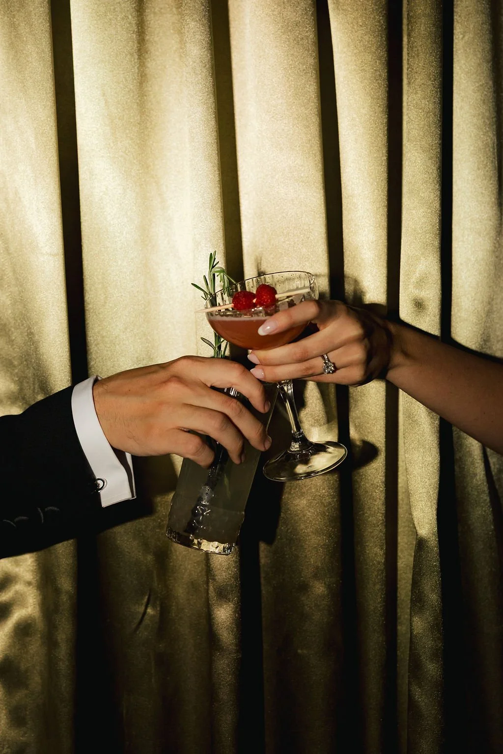 Two people holding cocktail glasses in a toast, with one person wearing a black suit and the other with a ring on their finger, against a gold curtain background.