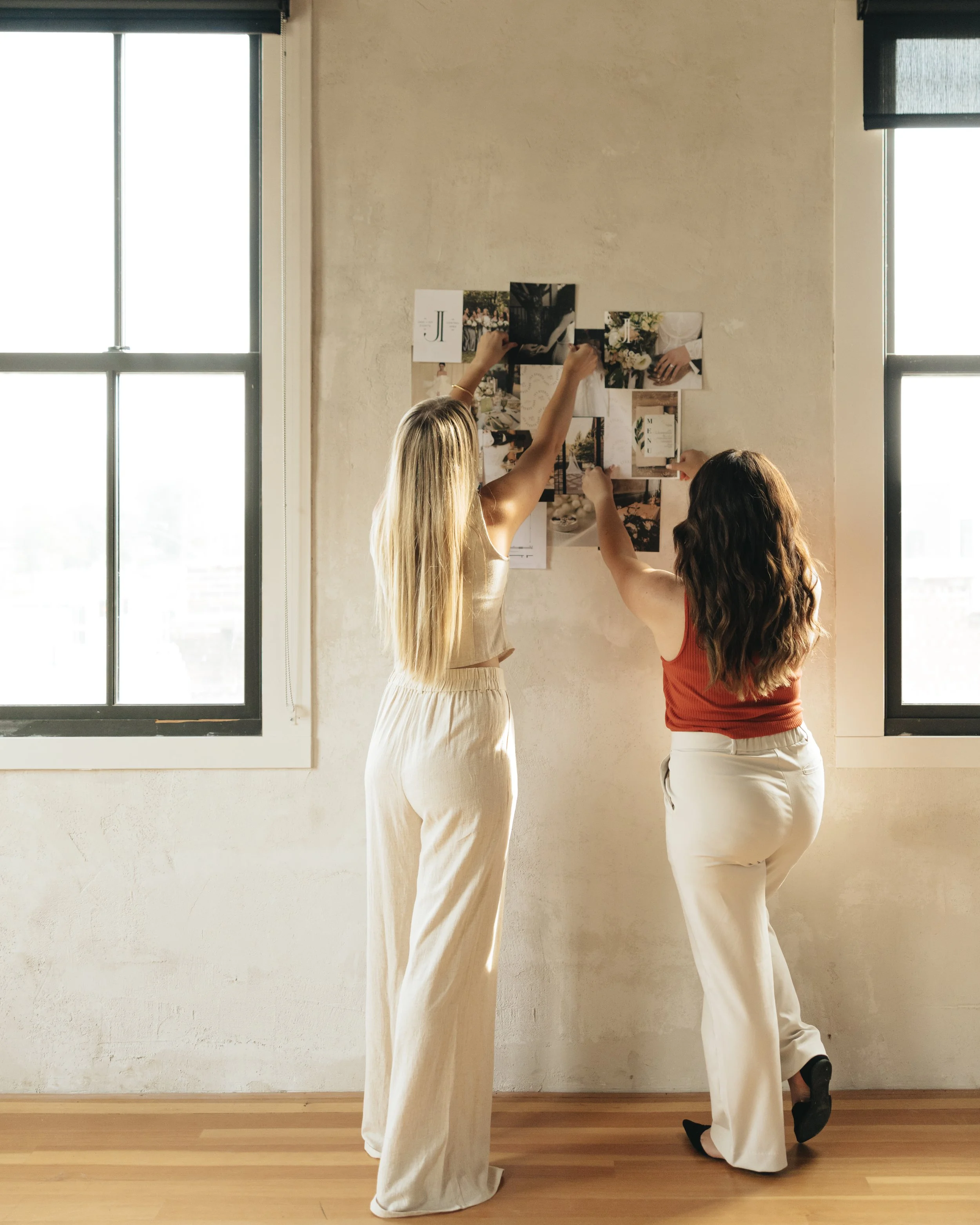 Two women with long hair reaching the wall, looking at and arranging photographs and prints on the wall, with large windows in the background.