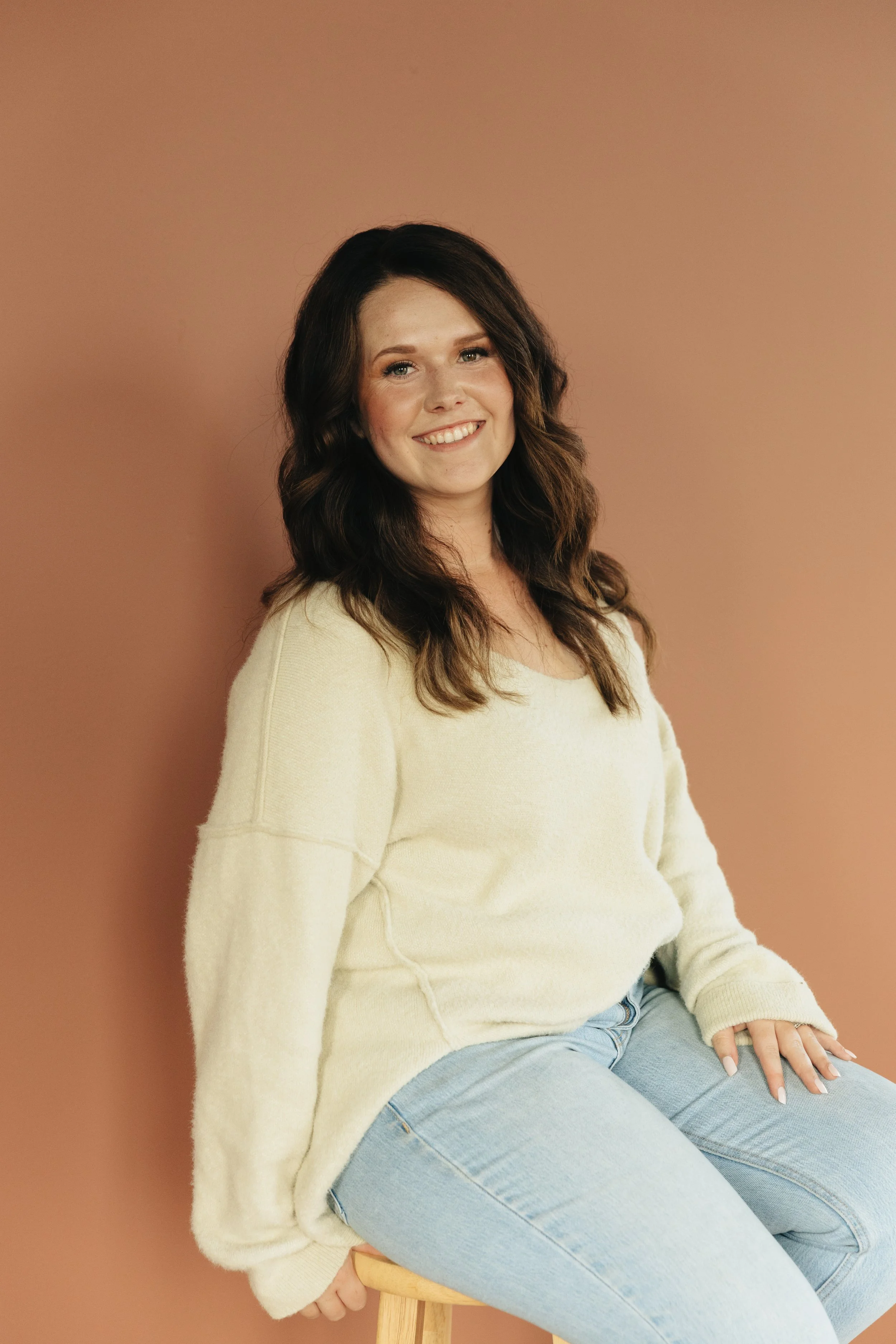 A woman sitting on a stool against a pink wall, wearing a cream-colored sweater and light blue jeans, smiling and looking at the camera.