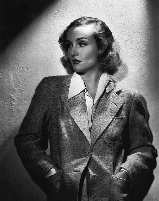 Black and white photo of a woman with vintage hairstyle and makeup, wearing a tailored blazer over a collared shirt, posing with hands in pockets against a textured wall.