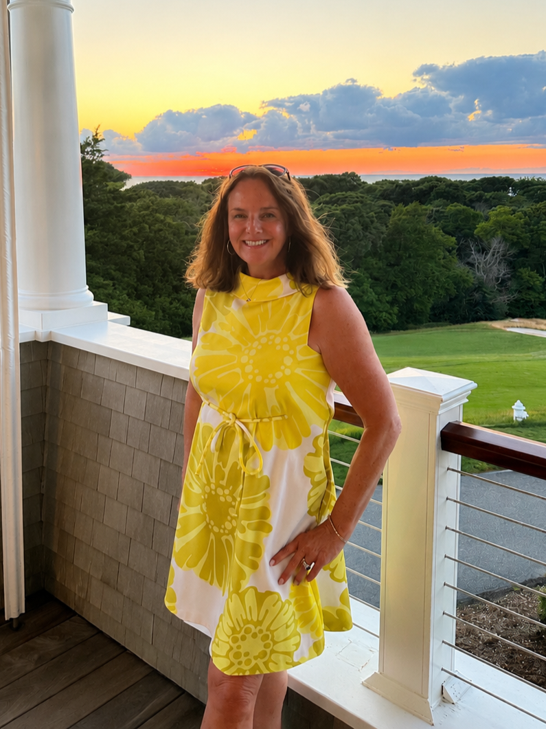 A woman in a yellow and white floral dress standing on a balcony at sunset with trees and a grassy area in the background.