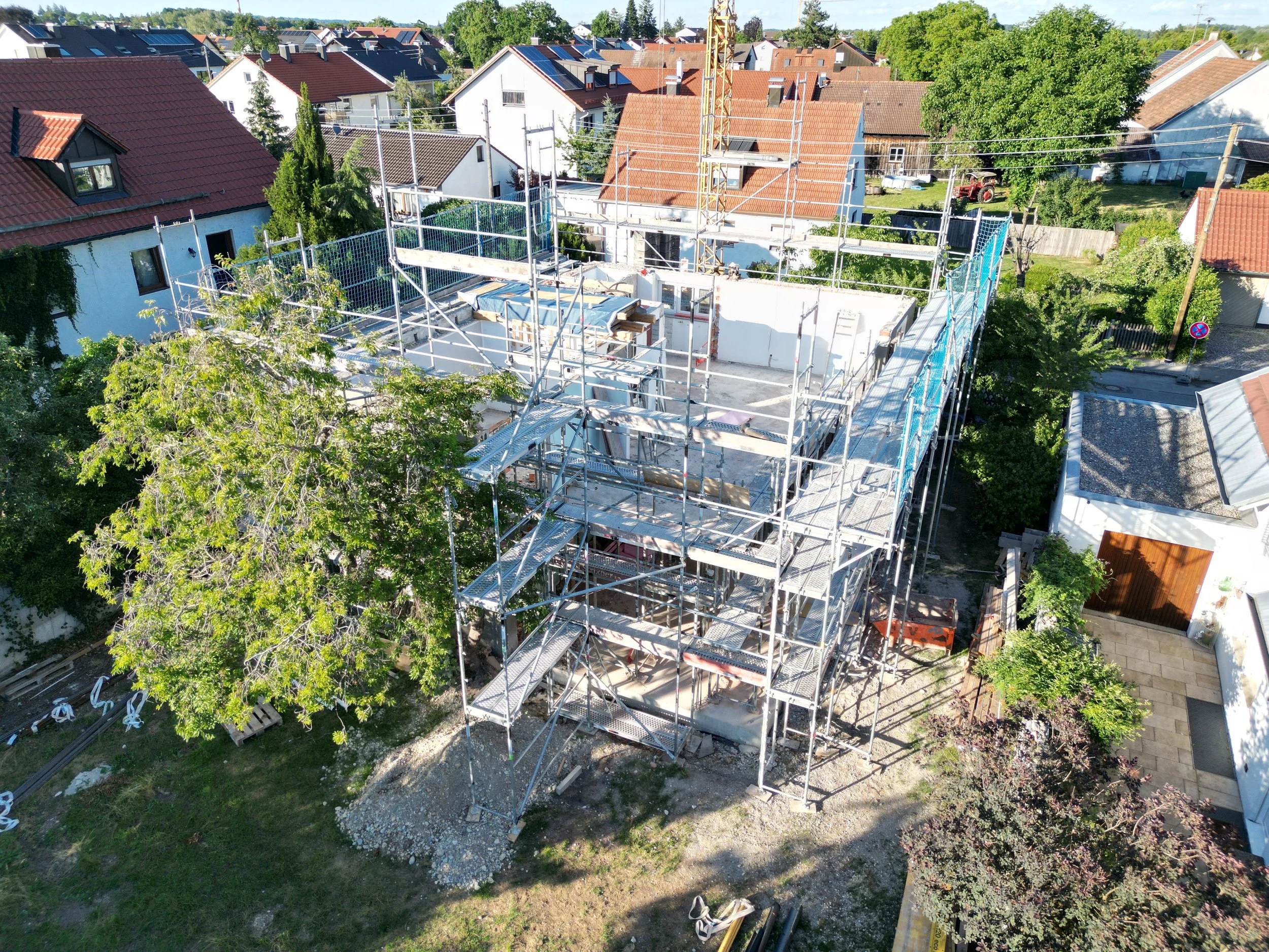Aerial view of a house under construction with scaffolding, surrounded by neighboring houses with red and dark roofs, trees, and clear sky.
