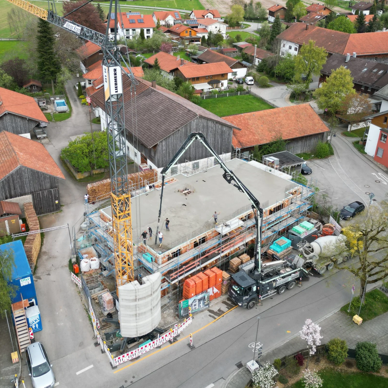 Aerial view of a construction site in a residential neighborhood, featuring a building under construction with concrete foundation, a crane, construction workers, and surrounding houses with red and brown roofs, trees, and parked cars.