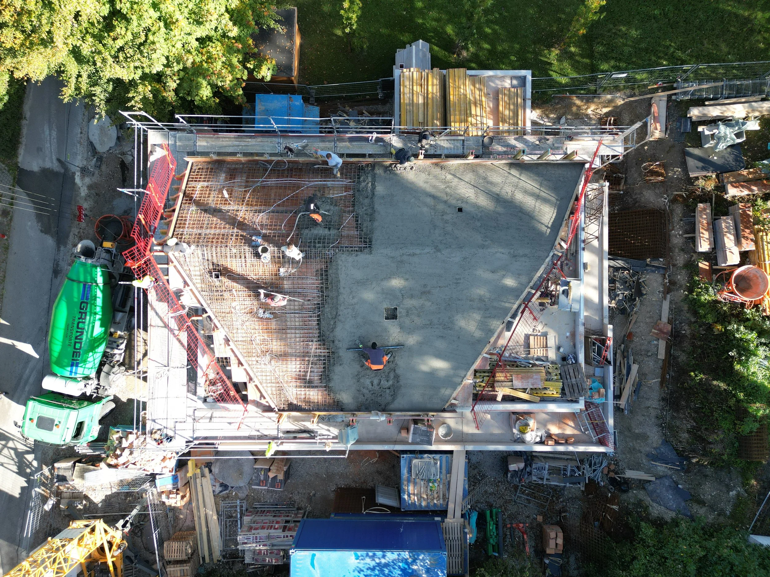 An aerial view of a construction site with workers, scaffolding, construction materials, and equipment, surrounded by trees.