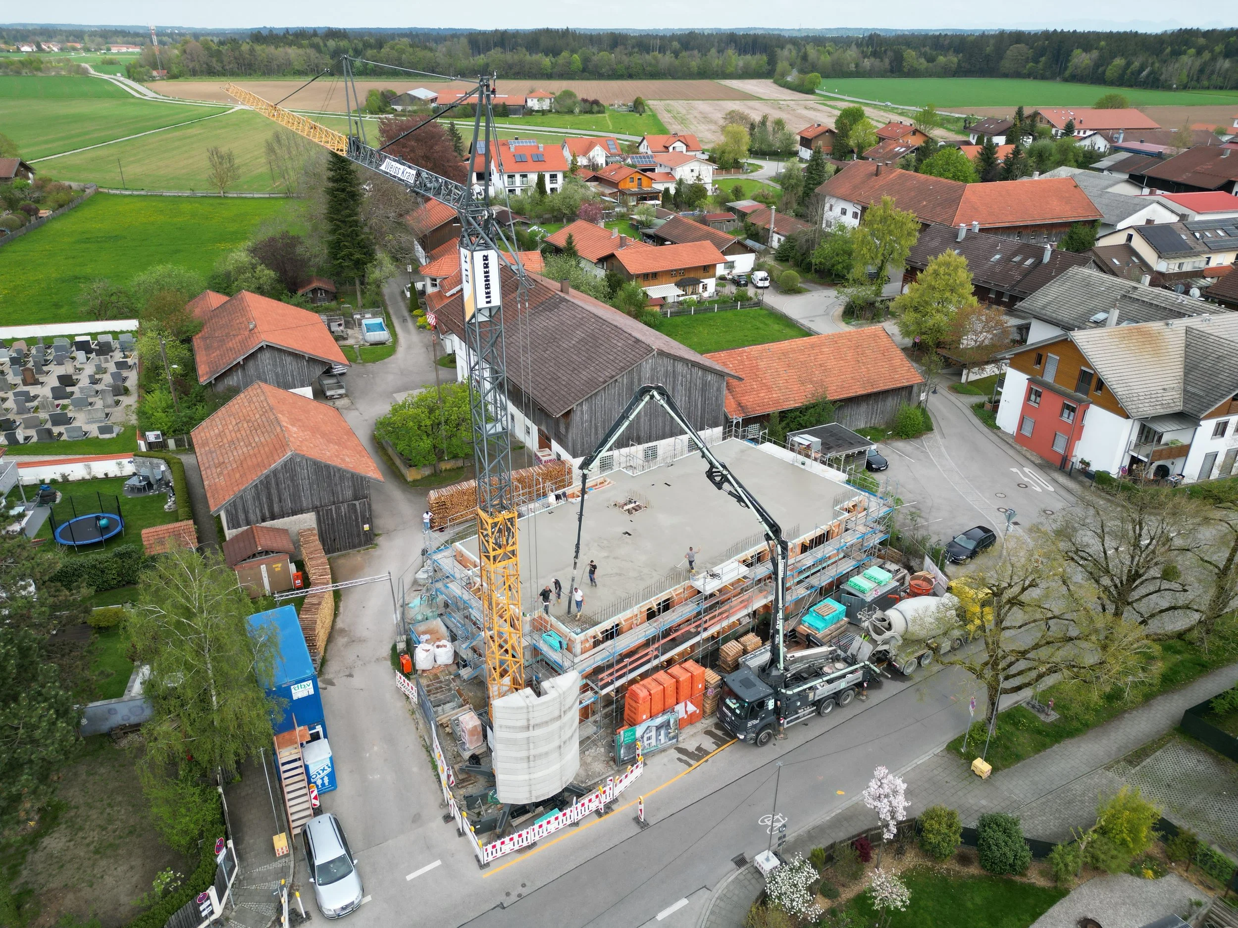 Ein Hausbau auf einer Baustelle in einer kleinen Stadt, mit Baukränen, Baugerüst und Baufahrzeugen, umgeben von Häusern und grünen Landschaften.