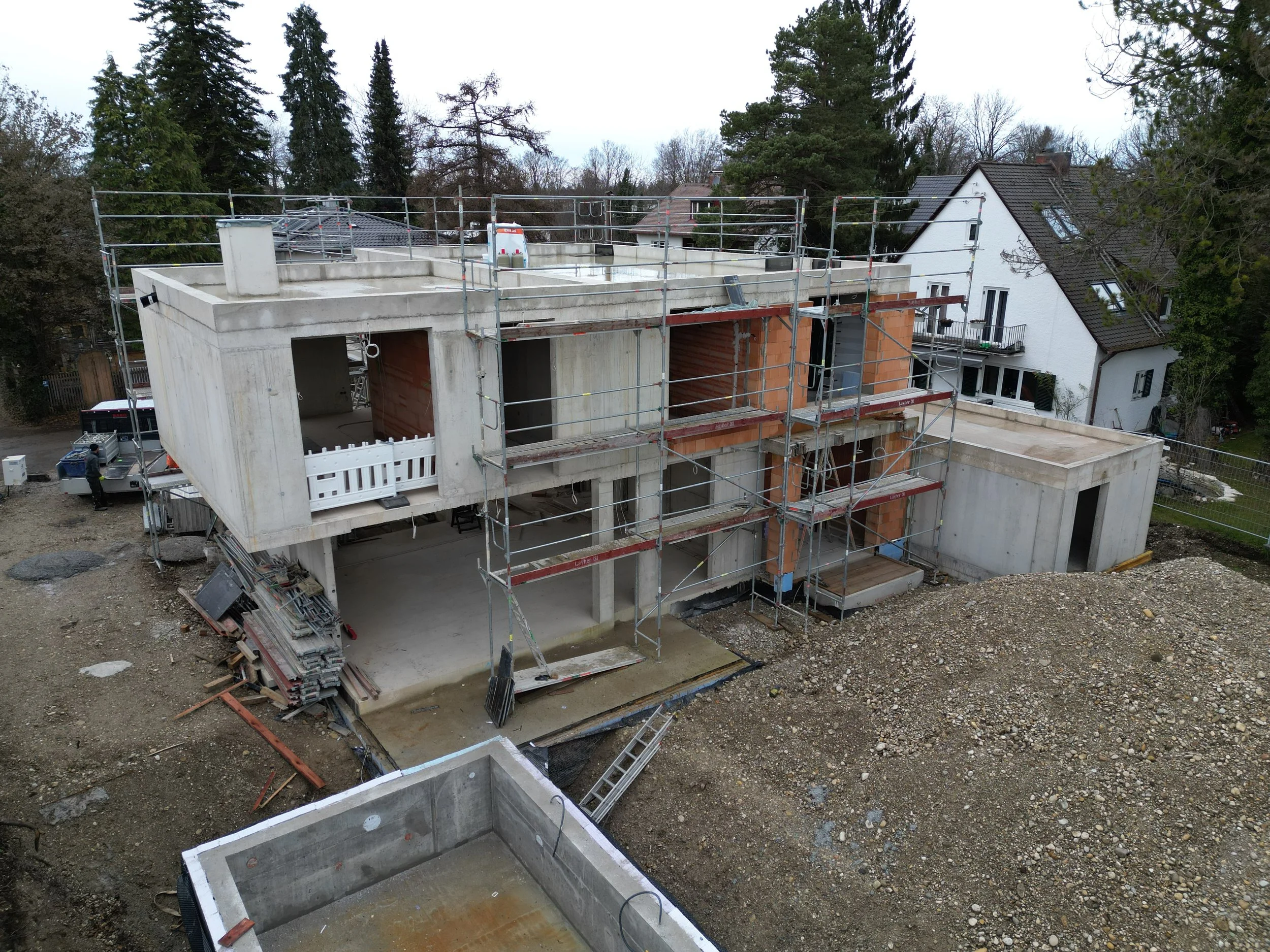 Construction site with a partially built house in urban residential area, scaffolding around the structure, trees, and neighboring houses visible in the background.