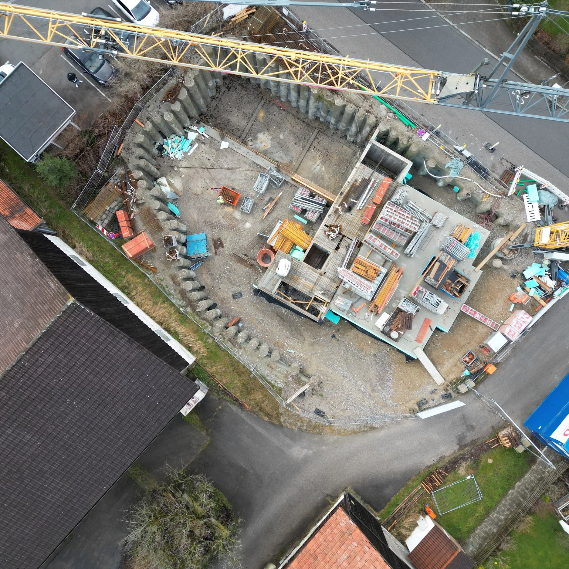 An aerial view of a construction site excavated in the ground, surrounded by a tall yellow crane, fences, and various construction materials and equipment. Nearby are neighboring buildings, trees, and paved roads.