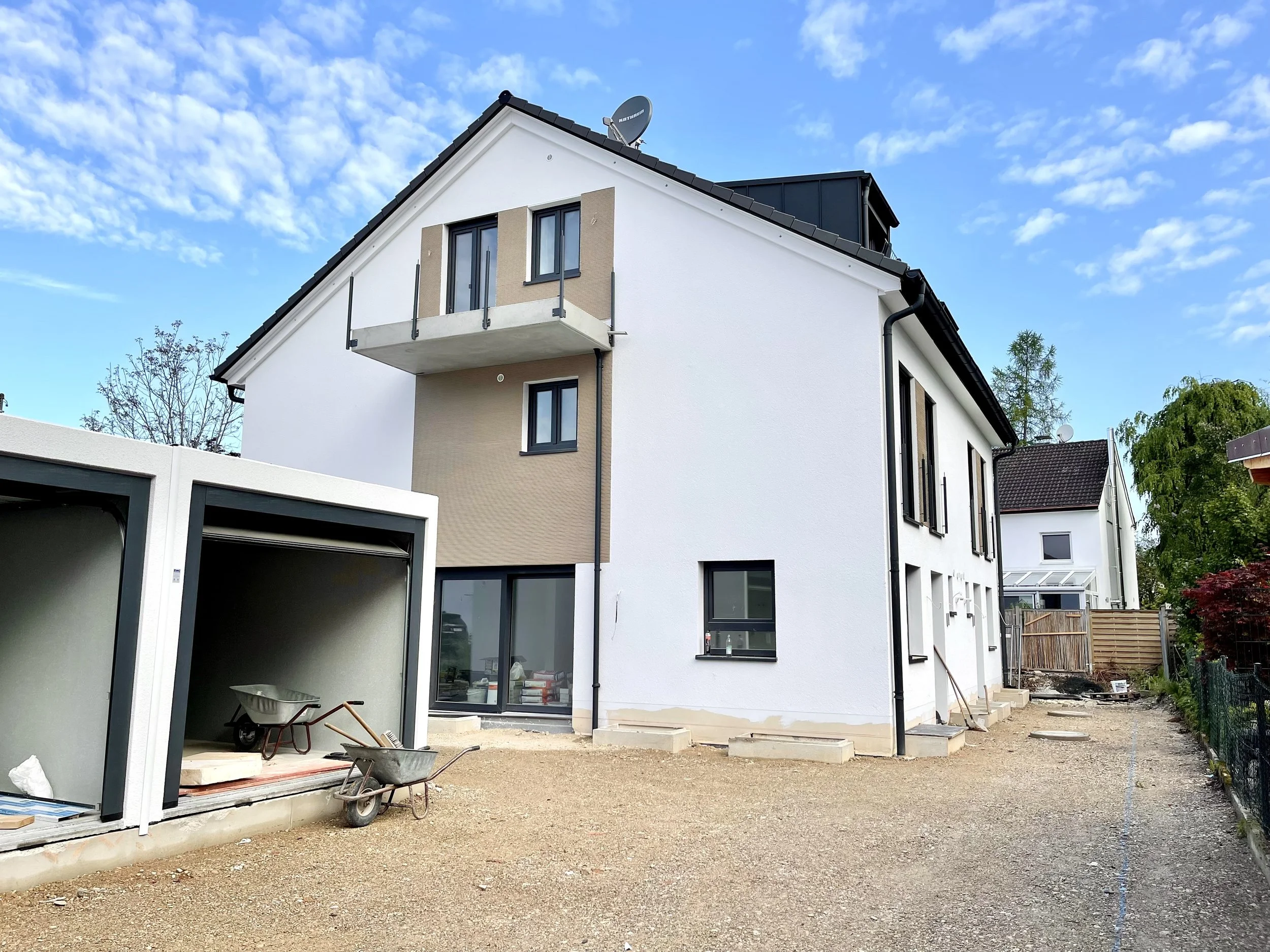 A modern multi-story house under construction with a white exterior and a small balcony on the second floor. There is construction material, including a wheelbarrow and tools, on the ground. The house has large windows, and the sky is partly cloudy.
